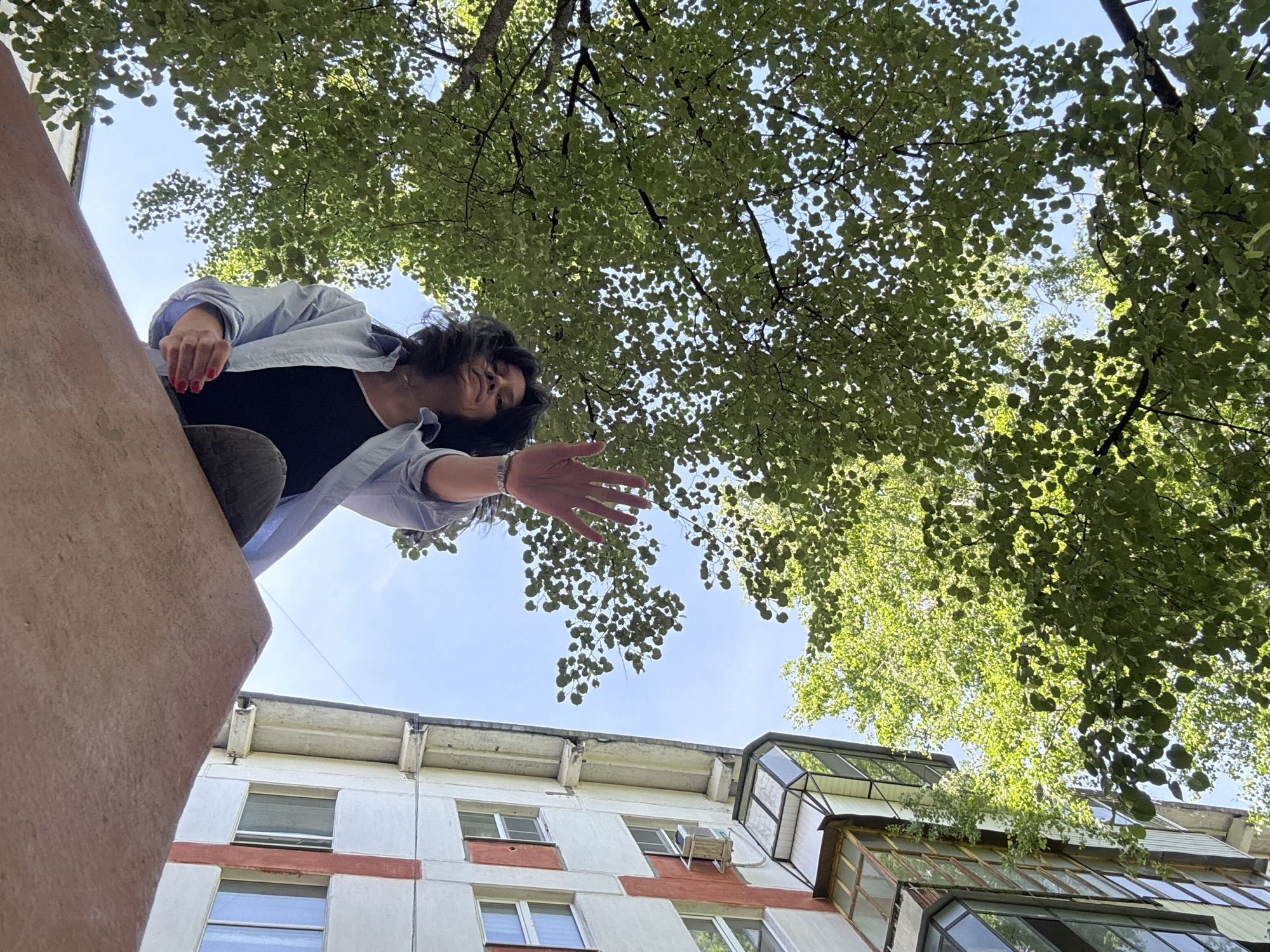 The image, shot from a low angle, features a woman partially visible in the upper left quadrant. She wears a light blue shirt over a dark top and has dark hair. She is reaching out with her right hand, wearing a bracelet on her wrist and red nail polish on her fingers.
Above her, the sky is partially obscured by dense green foliage from trees. In the bottom half of the frame, a multi-story building is visible. The building is painted white with red stripes between the windows, and it features a balcony with a glass enclosure. The sky is blue.
The location appears to be an urban setting.