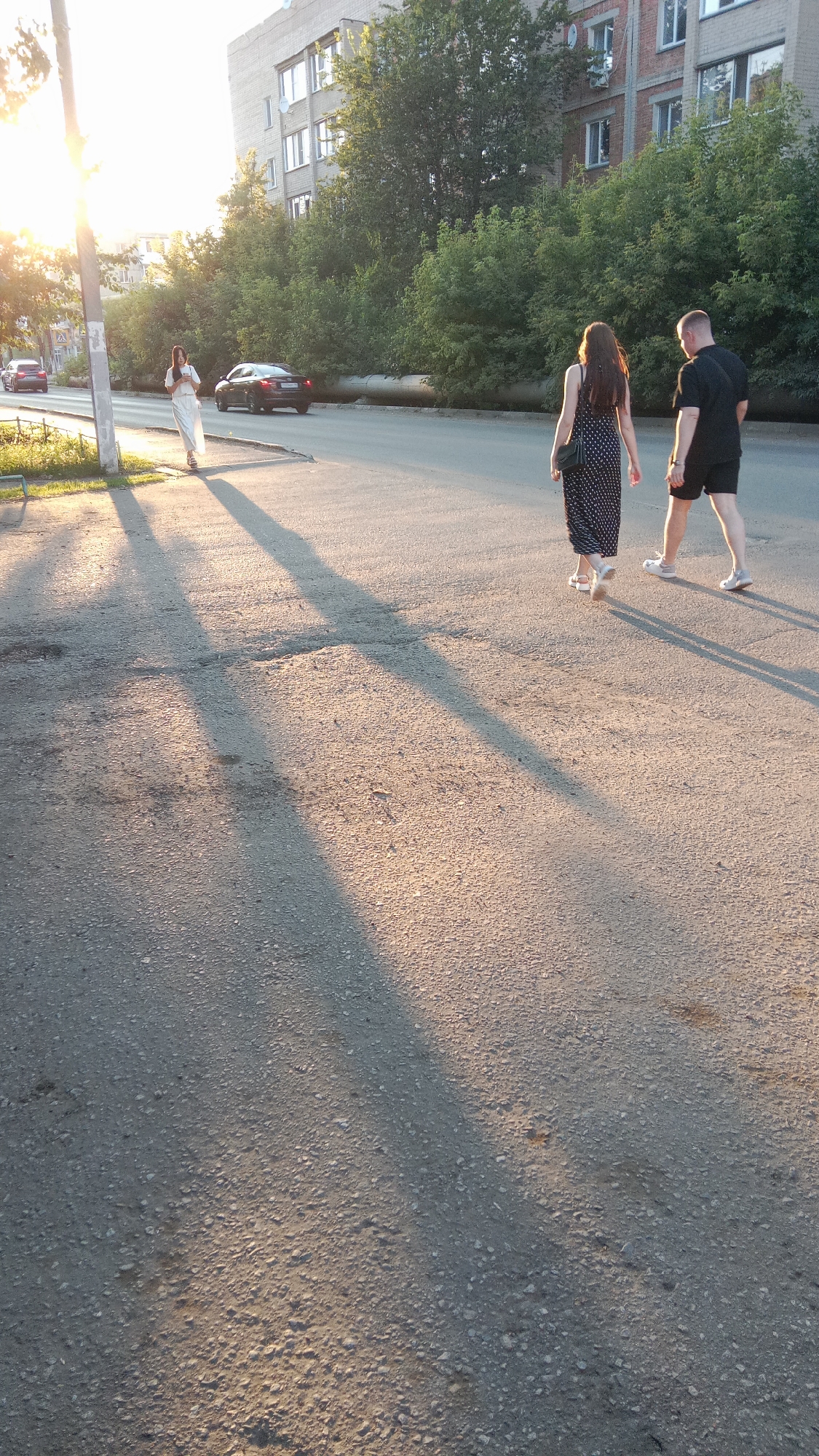 The image depicts an outdoor urban scene, likely in the city of Petropavl, Kazakhstan, during what appears to be late afternoon or early evening, given the long shadows cast by the low sun. The foreground is a grey asphalt road surface with visible cracks and scattered small stones. In the middle ground, a paved walkway or sidewalk runs parallel to the road.
On the walkway, three individuals are present. To the left, a person in a long, white flowing dress stands facing away from the camera, holding a phone in their right hand. The sun is positioned to the left and slightly behind this person, creating a bright lens flare effect and long shadows stretching across the road. Further down the walkway, a couple is walking away from the camera towards the right. The woman is wearing a black and white polka dot maxi dress and white sandals, carrying a black shoulder bag. The man is wearing a black t-shirt and black shorts with white athletic shoes.
In the background, a black sedan is driving on the road in the same direction as the couple. Beyond the walkway, a row of green trees and bushes forms a natural boundary. Behind the trees, a multi-story residential building with a grey facade and visible windows stands prominently. Another, shorter building with a brick facade is partially visible to the right of the grey building. A street light pole is situated on the left side of the image, near the person in the white dress. The overall environment suggests a residential or mixed-use urban area.