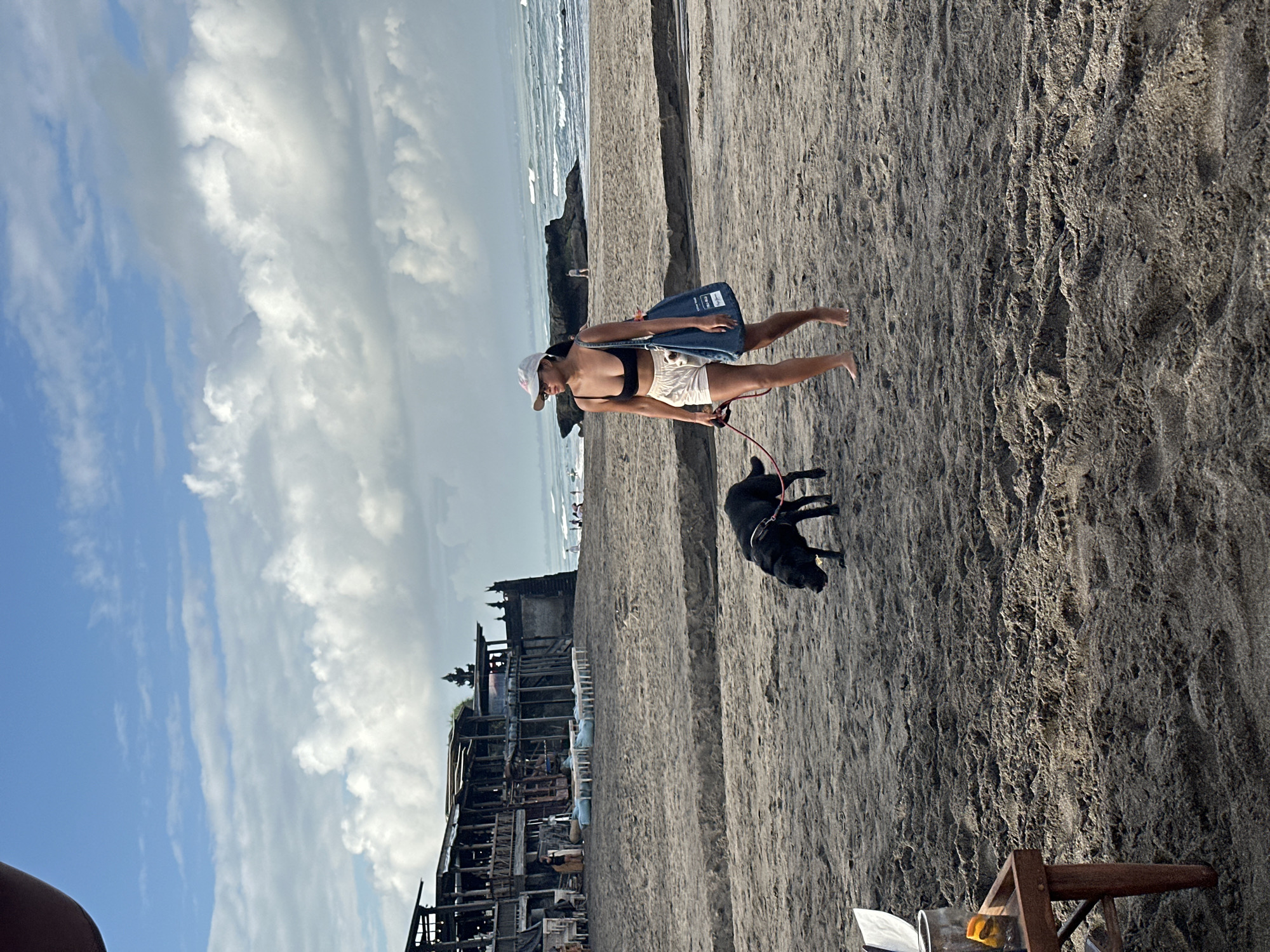 A woman is walking a black dog on a sandy beach in North Kuta, Indonesia. The woman is wearing a black bikini top, white shorts, and a white baseball cap. She has a blue tote bag slung over her shoulder. The dog is on a red leash. In the background, there is a multi-story wooden building with tables and chairs set up outside. The ocean is visible in the distance with waves breaking on the shore. The sky is partly cloudy. There is a small wooden table with a few items on it in the foreground to the left.