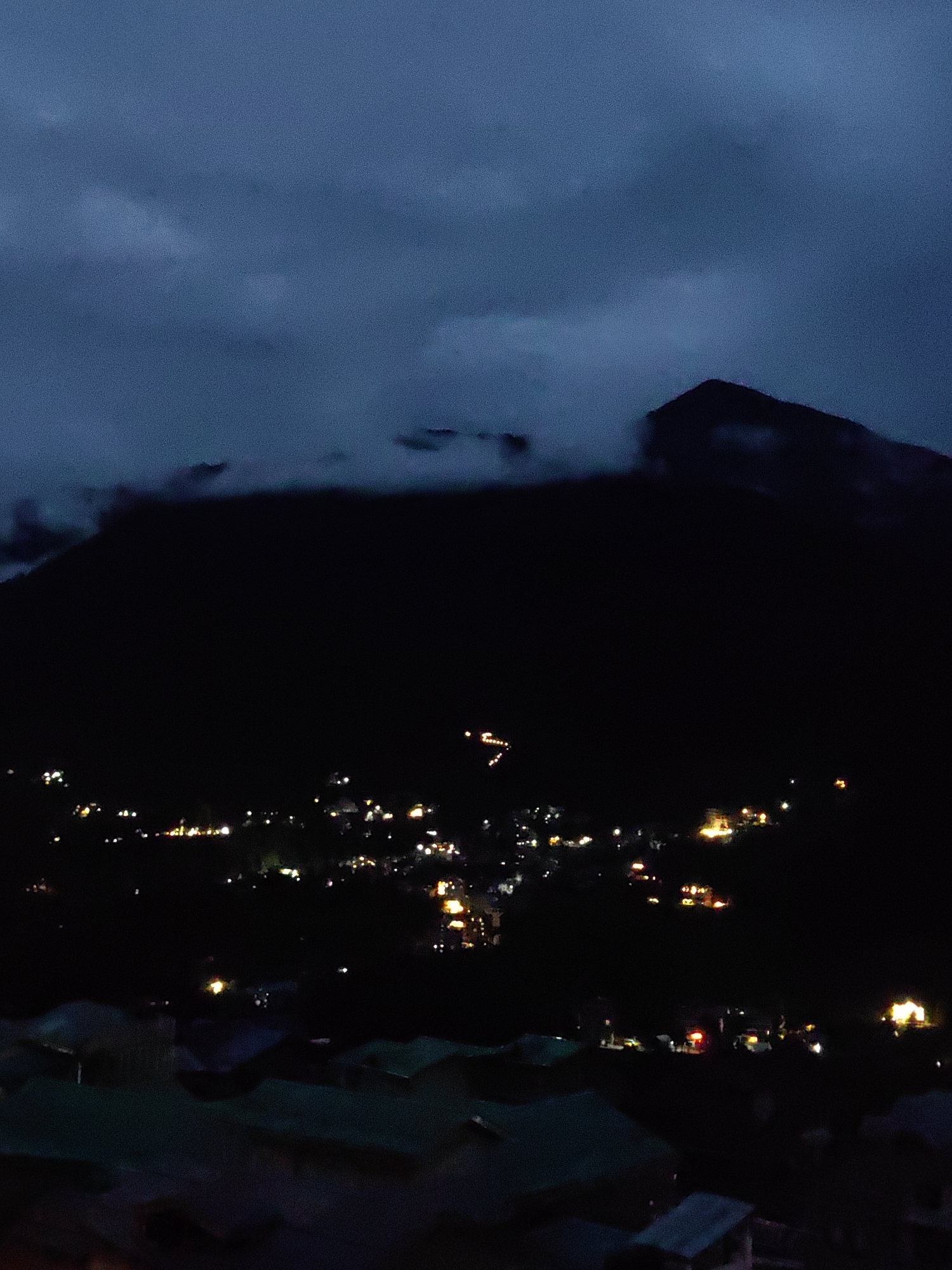 The image shows a nighttime view of Vashisht, India, with a mountain looming in the background. A dark, cloudy sky hangs over the scene. The town below is illuminated by scattered lights from buildings and streetlights, creating a warm glow against the darkness. The lights form a dense pattern, suggesting a populated area. The foreground shows the dark silhouettes of rooftops, indicating a lower elevation perspective. The overall impression is of a quiet, peaceful night in a mountain village.