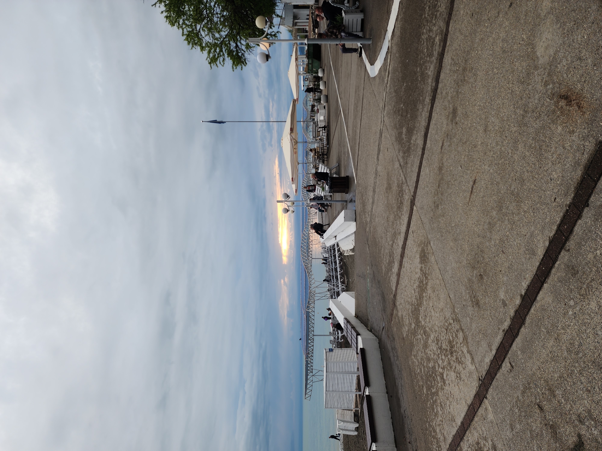 This image captures a serene evening view of a coastal promenade in Sochi, Russia, during sunset. The scene is oriented vertically but depicts a landscape along the waterfront.
On the right, a wide concrete pathway stretches into the distance, marked by darker embedded lines and a curved white painted line along its edge. To the left of the pathway, several outdoor seating areas, likely part of cafes or restaurants, are arranged. These include white lounge chairs, tables, and light-colored umbrellas, suggesting a relaxed atmosphere. Numerous individuals are visible, mostly seated and enjoying the evening by the water, engaging in activities such as relaxing, dining, or socializing.
Two multi-headed street lamps with spherical white globes illuminate sections of the promenade. A tall flagpole stands prominently in the middle ground, flying a dark blue flag against the sky. Further left, a series of white, latticed metal structures extend over the calm, light blue-green sea, possibly part of a pier or beach club infrastructure.
The horizon is dominated by a vibrant sunset, casting a golden glow through a band of clouds that stretch across the sky. The rest of the sky is overcast with soft, diffuse clouds. A tree with green foliage is visible in the upper left corner, adding a natural element to the urban-coastal setting. The overall mood is peaceful and leisurely, characteristic of a resort destination at dusk. No discernible text is visible.