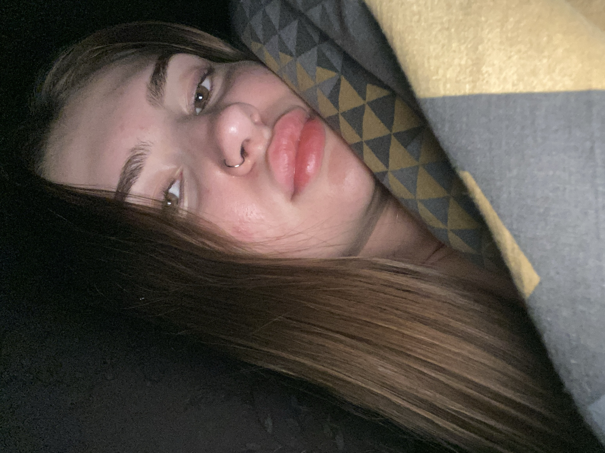 The image is a close-up, low-angle, portrait-style selfie of a young woman. She is partially covered by a blanket with a geometric pattern of grey and yellow triangles. Her head is tilted, and her gaze is directed upwards towards the camera. Her eyes are open, and she has plump, pink lips pursed slightly. She is wearing a nose ring. Her long, brown hair cascades down her left side, obscuring part of her face and the background. The lighting appears to be dim, suggesting it might be nighttime or an indoor setting with low light. The overall mood conveyed is relaxed, perhaps even sleepy or playful due to the expression and the way she is wrapped in the blanket. There are no visible text elements in the image. Given the location context of Дьяково, Russia, the setting is likely a bedroom or a similarly comfortable indoor space.