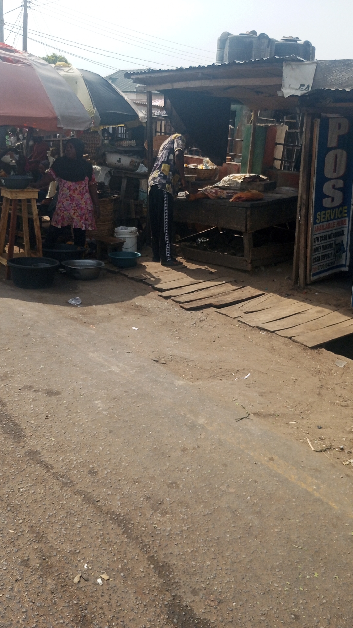 This image depicts a vibrant outdoor market scene in Lokoja, Nigeria, under a bright, sunny sky. The foreground consists of a gravelly and dirt ground, leading towards various market stalls constructed from wood and corrugated metal, some shaded by large, colorful umbrellas in red, white, and black. Overhead power lines crisscross the background against the clear sky.
Two main individuals are visible. On the left, a person in a floral dress and a dark head covering stands near a wooden stool and a black basin. On the right, another person, seen from behind in a patterned shirt and dark pants, is actively tending to items displayed on a wooden counter, which appear to be various food products. Other figures are faintly discernible in the background, suggesting a busy market environment.
Numerous objects populate the scene, including several basins and buckets (black, silver, blue, white) on the ground near the stalls. A makeshift wooden walkway, made of planks, bridges a small ditch or gutter in front of the rightmost stall. Two large black water tanks are visible on the roof of one of the market structures.
A prominent blue sign on the right advertises "POS SERVICE," detailing financial services available. The visible text includes "SERVICE AVAILABLE HERE," "ATM CASH WITHDRAWAL," "MONEY TRANSFER," "CASH DEPOSIT," and a contact number, "TEL: 070311542978." The strong shadows indicate it is daytime, likely mid-morning or early afternoon, with clear weather. The scene portrays a typical informal trading atmosphere.