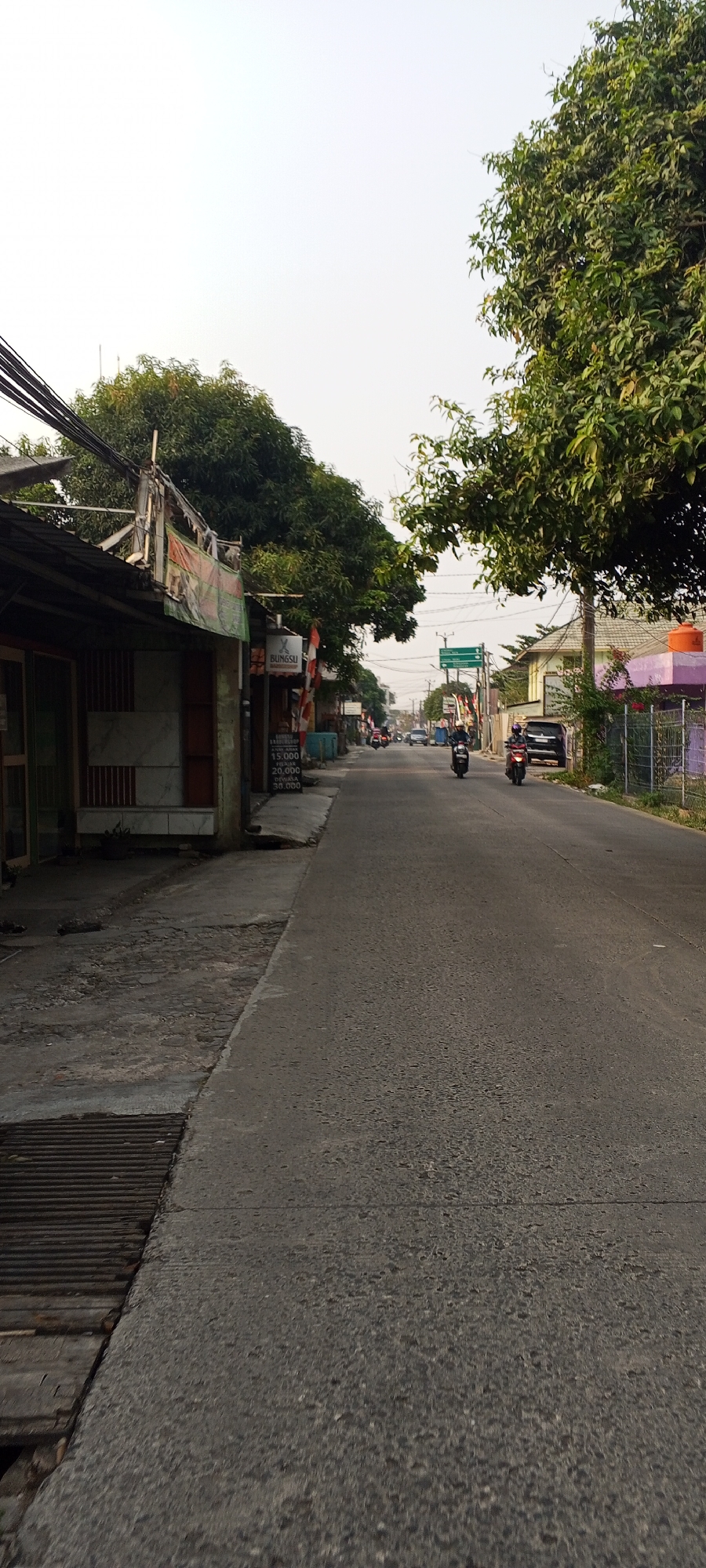 The image shows a street scene in Depok, Indonesia, likely in a residential or commercial area. A paved road stretches into the distance, lined on the left by low-rise buildings with awnings and on the right by houses and trees. Power lines are visible overhead. Several motorcycles are present, with two in the foreground and more further down the road. One motorcycle is parked on the side of the road near a building with a sign that reads "BUNGSU". A black car is also parked on the right side of the road. The sky is bright and overcast. The overall impression is of a typical urban street on a cloudy day.