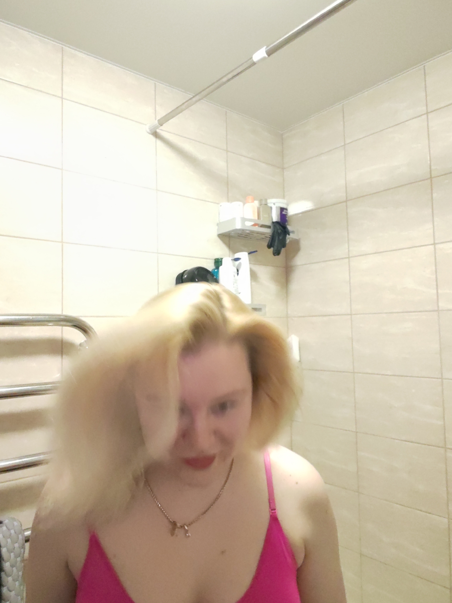 A woman with blonde hair, wearing a pink top and a gold necklace, is in a tiled bathroom. Behind her is a towel warmer and a shelf with toiletries. The location is likely Izhevsk, Russia.