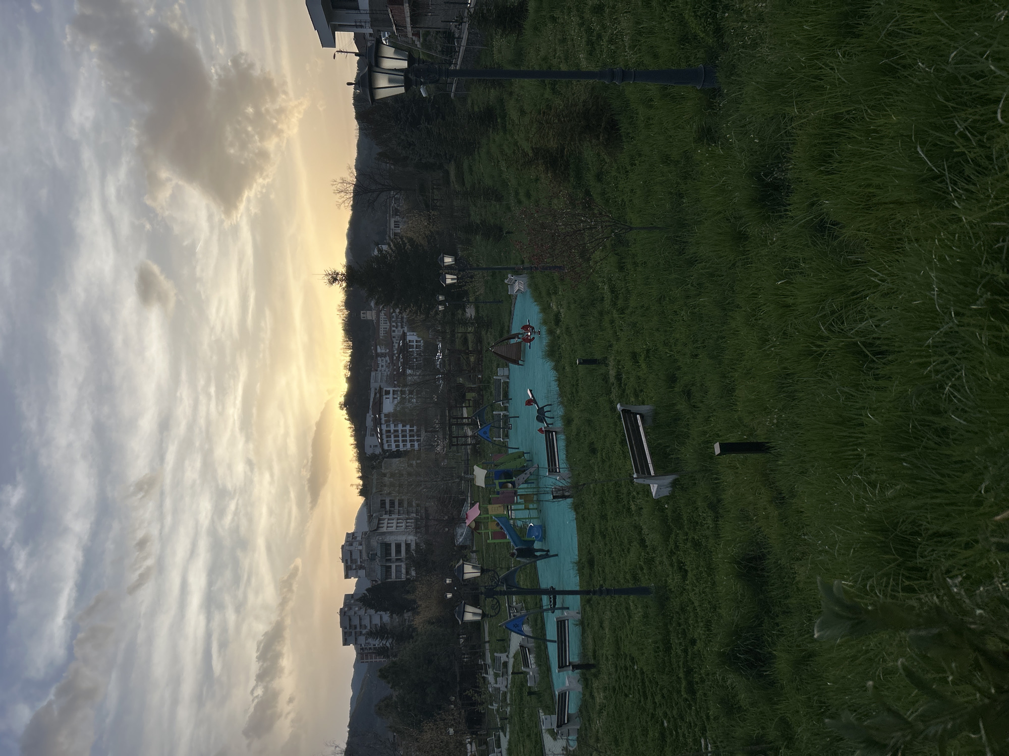 The image shows a park or green space in Zlatograd, Bulgaria, during the late afternoon or early evening. The sky is filled with clouds, with a soft, golden light indicating the proximity to sunset. A hill or mountain range is visible in the distance.
In the foreground, there is a grassy area, well-maintained, leading to a playground with various play structures. A blue rubber surface is installed around the playground equipment. Benches and lampposts line the park's pathways. The architecture of the buildings in the background indicates an urban environment. No people are visible in the scene.