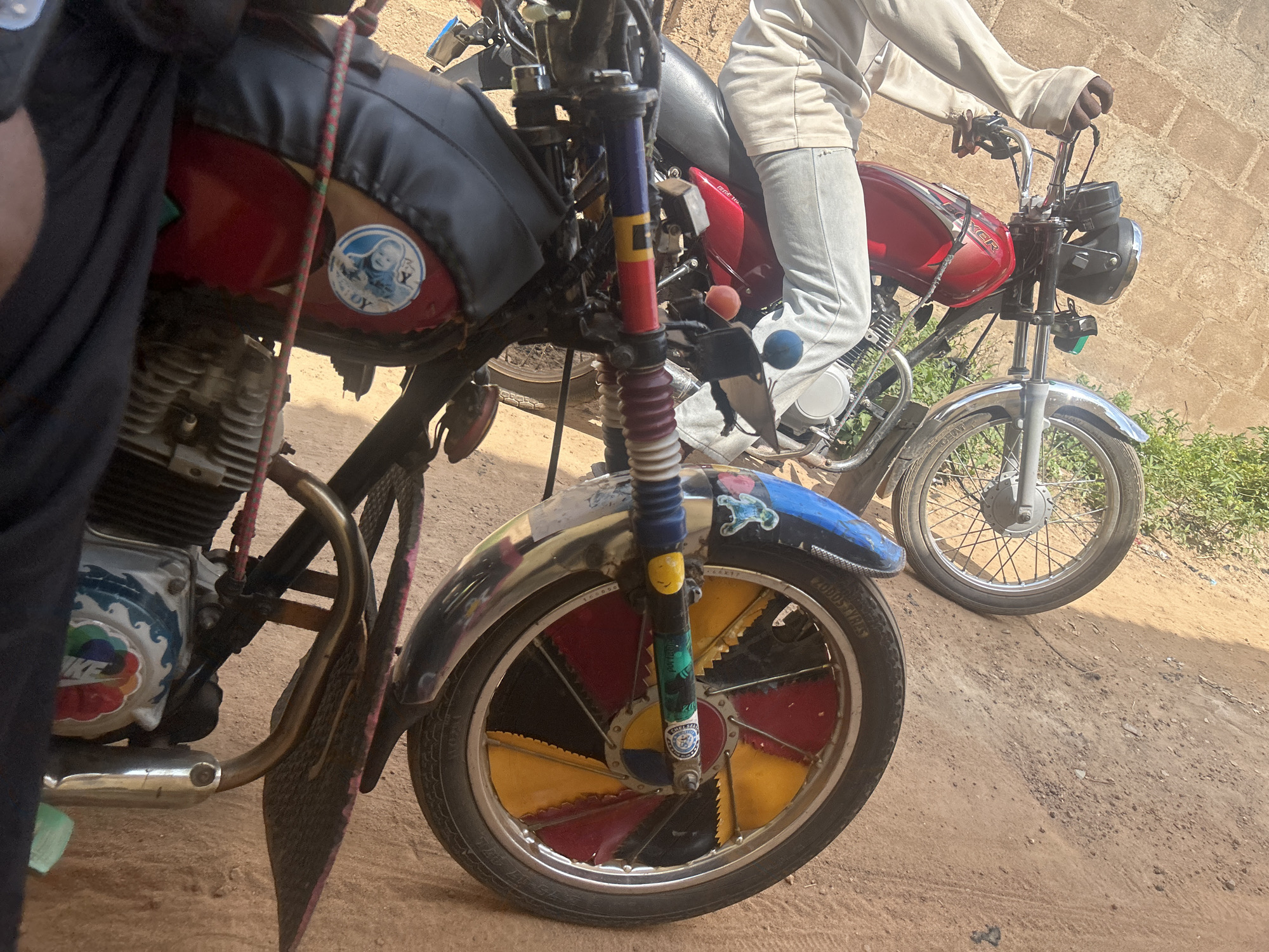 The image shows two motorcycles parked side-by-side on a dirt road. The foreground motorcycle is red and black, with a person's legs visible on the right side. It has a decorative wheel with red, yellow, and black sections. The motorcycle also features stickers on its fuel tank, one of which depicts a baby. The handlebars are colorful, with red, blue, and yellow accents.
Behind the foreground motorcycle is another red motorcycle, on which a person is seated. This person is wearing light-colored pants and a long-sleeved shirt. The background shows a brick wall and some greenery, suggesting an outdoor setting. The lighting indicates it is daytime, with shadows cast by the motorcycles. The overall scene appears to be in Ogbomosho, Nigeria, with the dirt road and brick wall being characteristic of the location. There is no discernible text in the image.