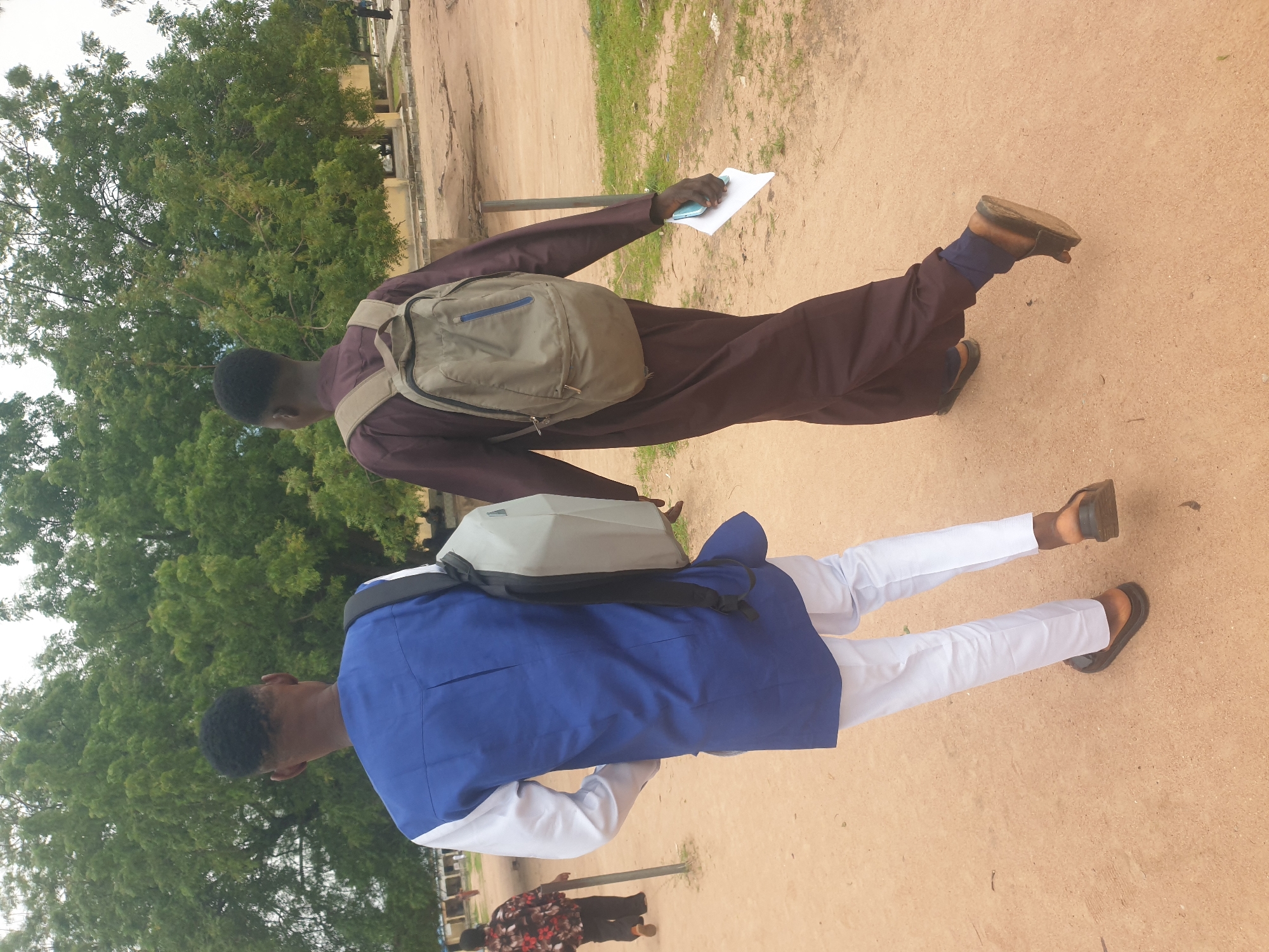 Two young men are walking away from the camera on a sandy path in an outdoor setting. The man on the left is wearing a blue tunic with white sleeves and white pants, and a grey backpack is slung over his shoulders. The man on the right is wearing a brown robe and also has a grey backpack. He is holding a white piece of paper in his right hand. In the background, lush green trees are visible, and a building with a light-colored facade is situated further back. The ground is a mix of sand and patchy grass. The lighting suggests it is daytime, possibly overcast.