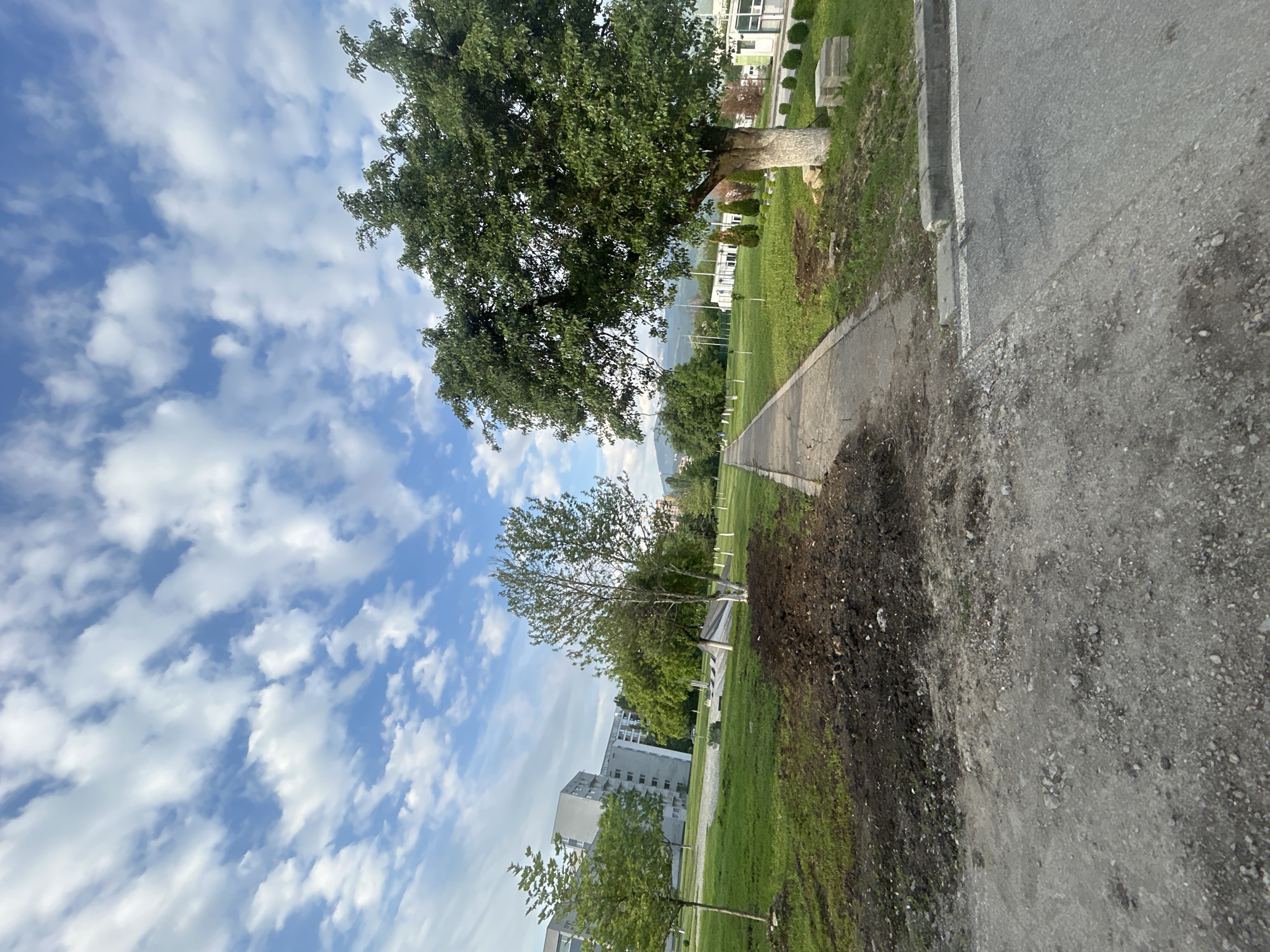 The image shows a scene in the city of Unknown, Unknown. The photo captures a partially clouded sky with several trees bordering a paved area. To the right of the paved area, there is a strip of grass with freshly disturbed soil along the edge. Buildings are visible in the distance, suggesting an urban or suburban setting. There are no people visible in the scene.