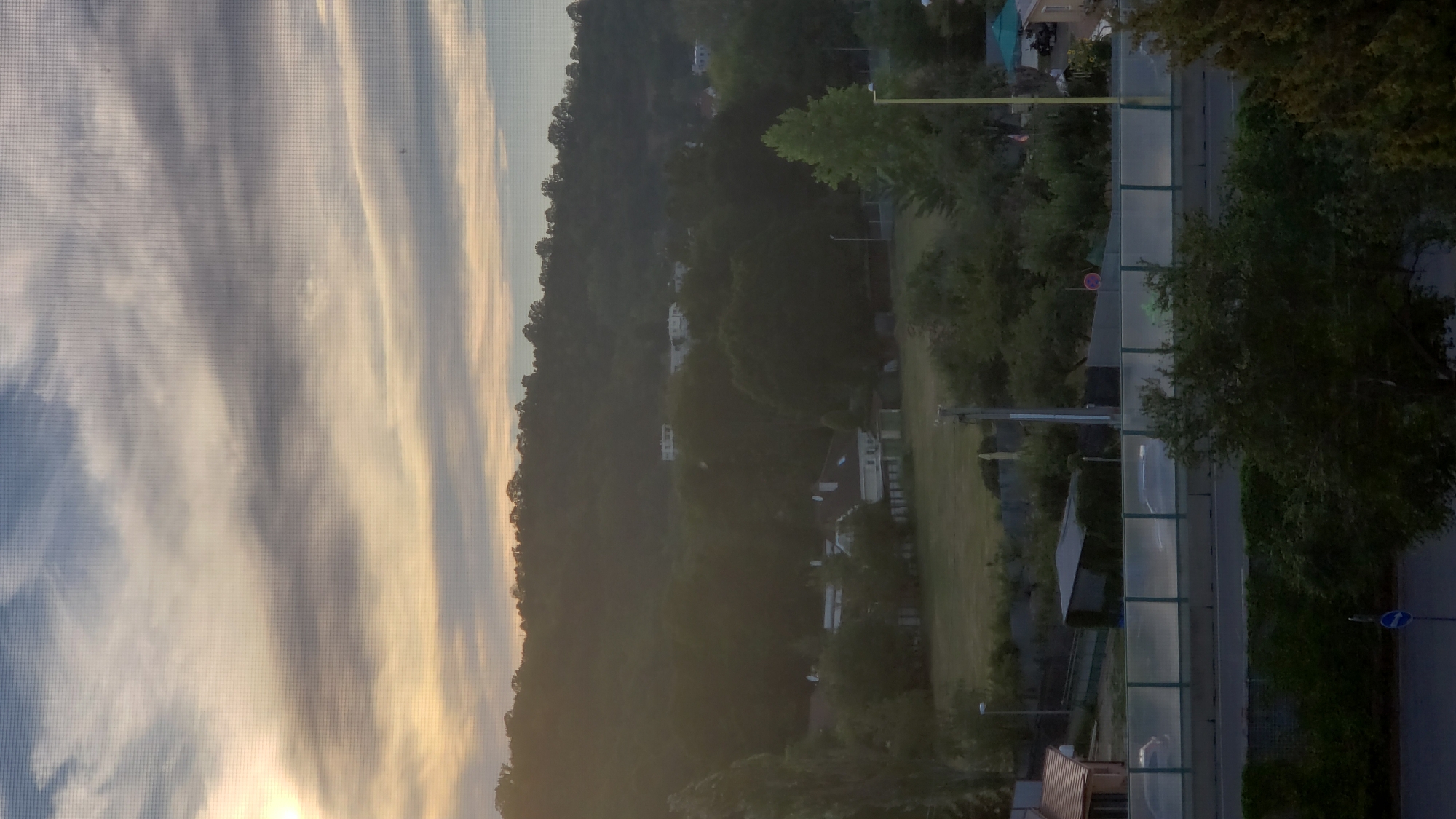 This is a photograph taken from a window with a screen, looking out at a suburban landscape during dusk. The sky is a mix of blue and grey clouds, with the sun setting in the upper left corner, casting a warm glow. In the foreground, a road is visible with a few trees and a traffic sign. Behind the road, there is a noise barrier, and beyond that, a grassy field with some buildings and more trees. In the background, a tree-covered hill rises, with a few houses or apartment buildings visible on its slope. The overall scene depicts a peaceful, residential area.