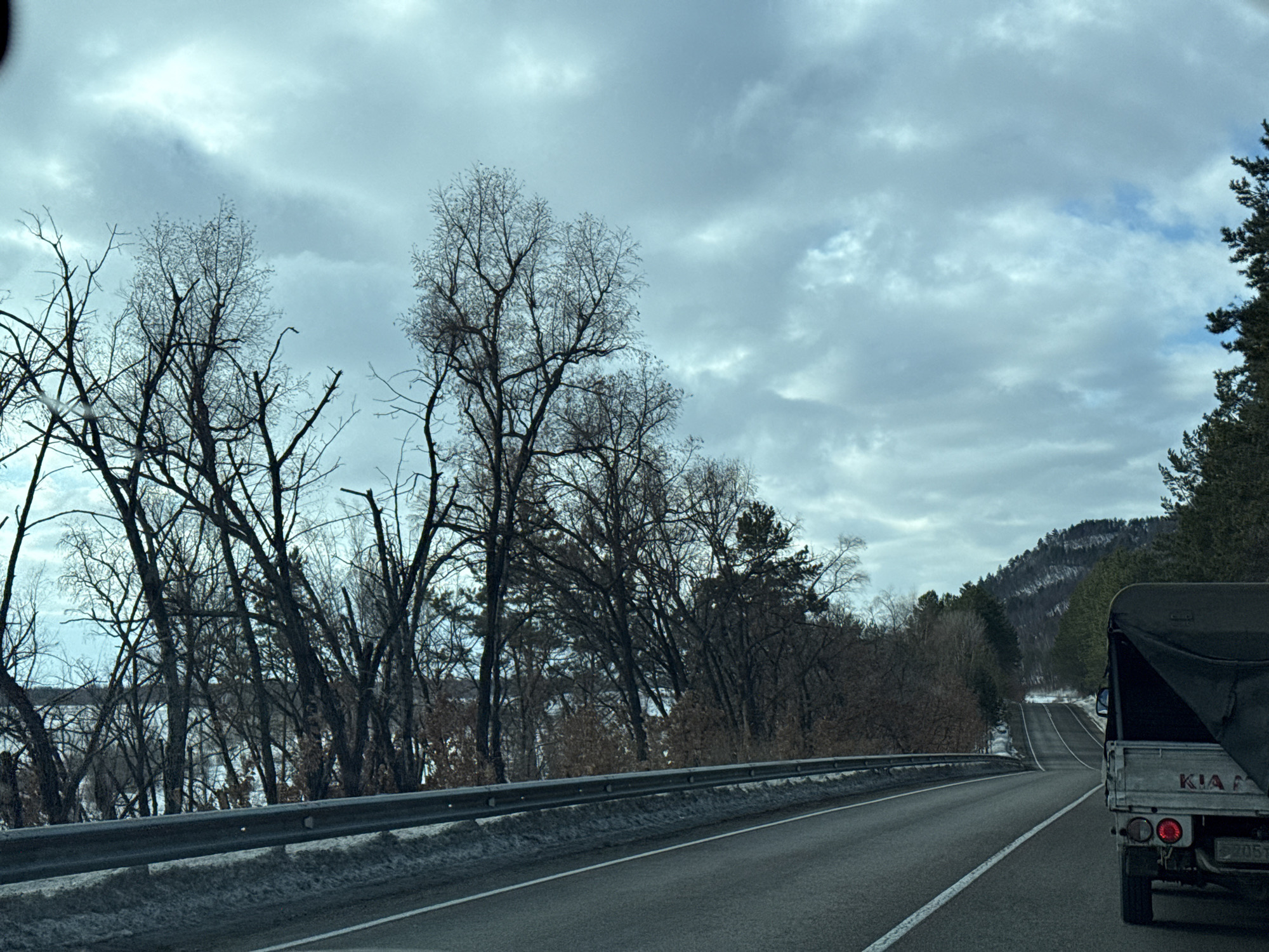An outdoor, forward-facing vehicle perspective captures a two-lane asphalt road under an overcast sky on a winter or late autumn day. A metal guardrail extends along the left side of the road, behind which a dense line of bare deciduous trees partially obscures a body of water, possibly frozen or ice-covered. In the right foreground, the rear of a light-colored KIA pickup truck, featuring a dark, tarp-covered bed, is visible; its license plate partially shows "2051", and it has circular taillights. The road ahead curves gently and ascends into the distance, bordered by mixed coniferous and deciduous trees leading to forested hills with visible snow patches. The sky overhead displays varying shades of grey and hints of blue. Traces of snow and ice are present on the road shoulders. The scene is located in Unknown, Unknown.