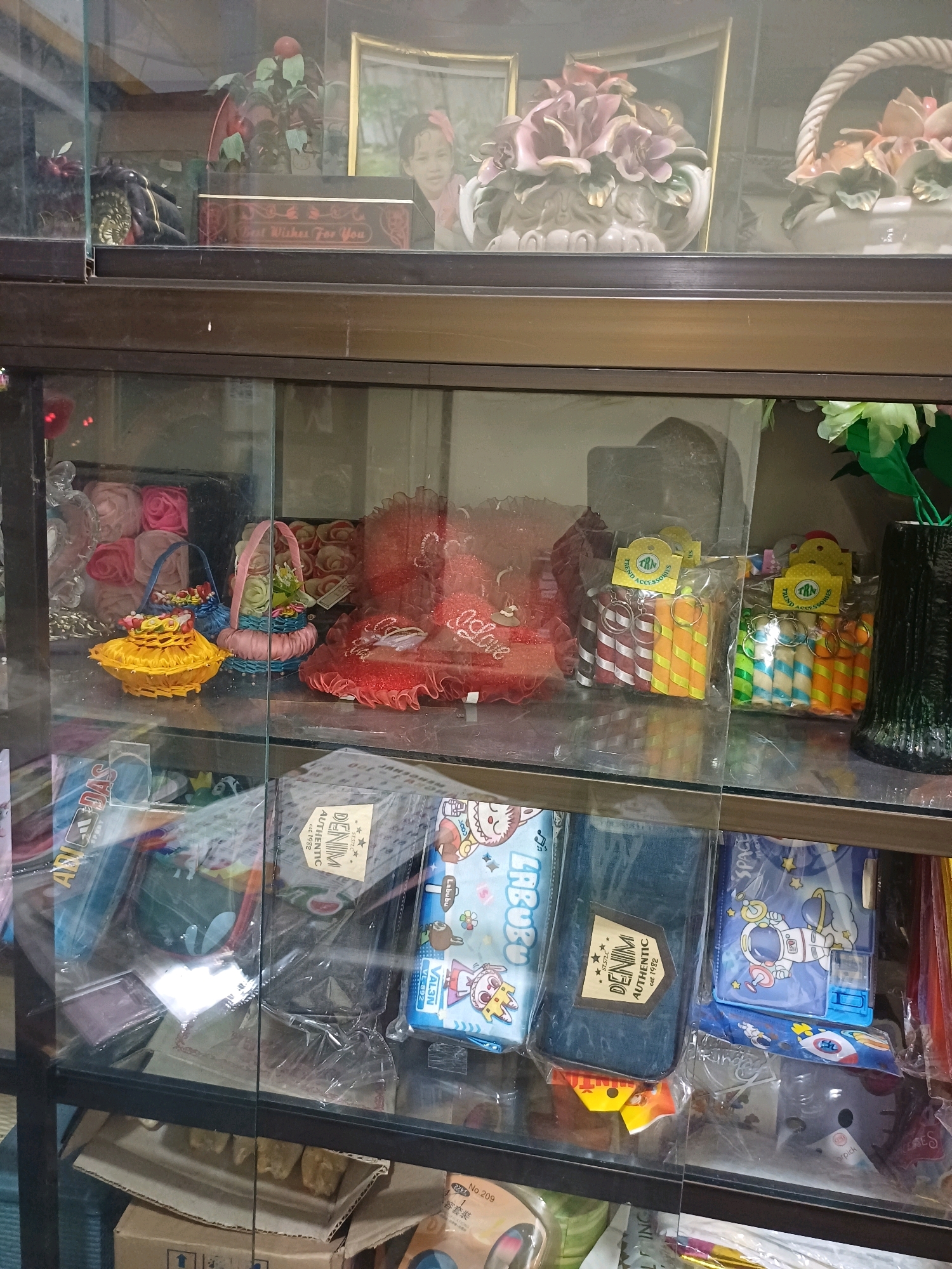 This is a medium-shot, eye-level photograph of items displayed in a glass-fronted cabinet, likely a shop or market stall in Dumai City, Indonesia. The cabinet has multiple shelves, and the contents are visible through the glass, which reflects some light.
The top shelf displays decorative items, including a framed picture of a child, ceramic flower arrangements, and a red heart-shaped object with the word "Love" on it. There are also some smaller baskets with colorful flowers.
The middle shelf features a variety of small, packaged items. There are two yellow baskets with colorful fabric flowers. To the right of these baskets are several small, individually packaged candies or candies that appear to be cylindrical and striped in various colors (red and white, orange and white, green and white, blue and white). These are likely novelty candies or party favors. Further to the right, there's a tall, dark, textured vase.
The bottom shelf contains an assortment of flat, packaged items. Some appear to be pencil cases or stationery pouches with colorful cartoon characters and patterns, including astronauts and rabbits. There are also what appear to be fabric or denim-like pouches with the label "DENIM AUTHENTIC est. 1982." Cardboard boxes are visible at the bottom of the cabinet.
The overall environment appears to be a retail setting, possibly a small shop or a stall in a market, focused on gifts, stationery, or novelty items. The lighting is somewhat dim, creating reflections on the glass. There are no people visible in the photograph.
