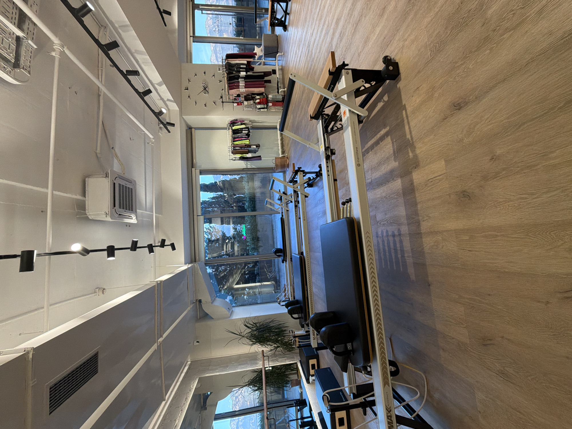 This image depicts the interior of a modern fitness studio, likely a Pilates studio, located in Boreti, Montenegro. The studio features a polished wooden floor and large windows that offer a view of a scenic mountainous landscape with some trees.
Dominating the foreground and midground are several Pilates reformers, which are exercise machines designed for resistance training. These reformers are predominantly white and black with padded carriages and footbars. Some appear to be Merrithew branded. To the left, there are additional black padded boxes used for Pilates exercises.
In the background, beyond the reformers, a clothing rack displays a variety of workout apparel, suggesting the studio also sells merchandise. A wall clock in an artistic bird design is visible on the right-hand wall. The ceiling is equipped with industrial lighting fixtures and what appears to be an air conditioning unit. The overall ambiance suggests a clean, well-lit, and well-equipped space for physical training. There are no people visible in the image.