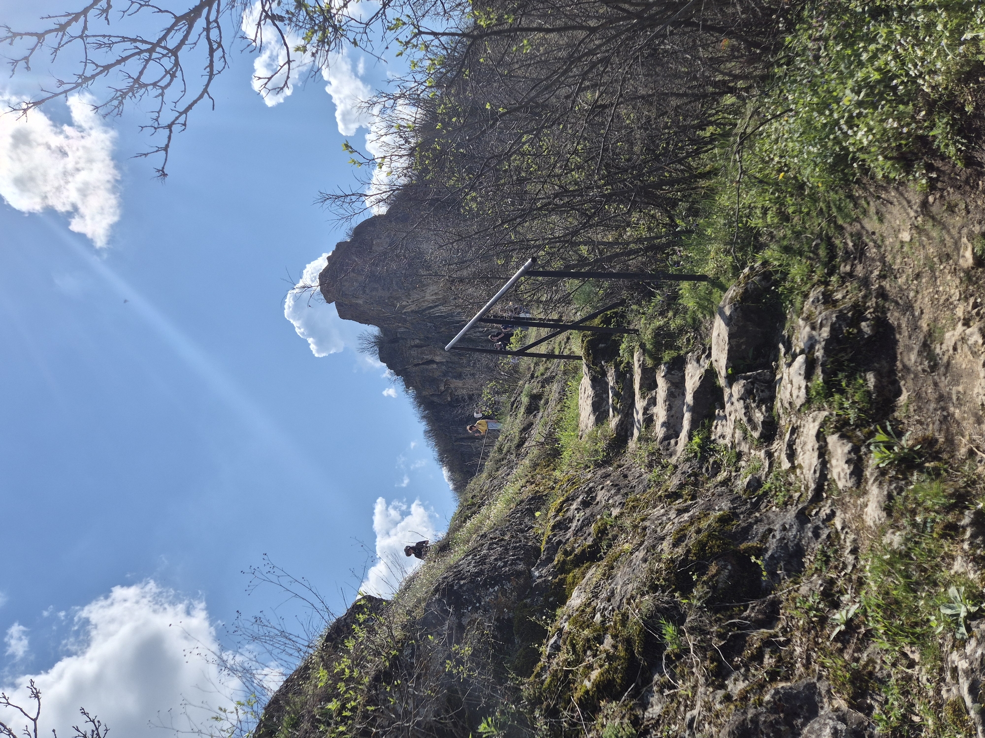 The image depicts a rocky uphill path with stone steps, possibly a trail, in a natural setting. There's a metal handrail on the path's left side. The terrain is rugged, with moss and vegetation growing on the rocks. In the background, a distinct rock formation protrudes against a blue sky with scattered clouds. Two people are discernible further up the path, one wearing a yellow shirt. The presence of trees and bushes suggests a forested area or hillside. This could be situated in Ivanovo, Bulgaria, but without further information, definitive localization to this specific area is not possible.