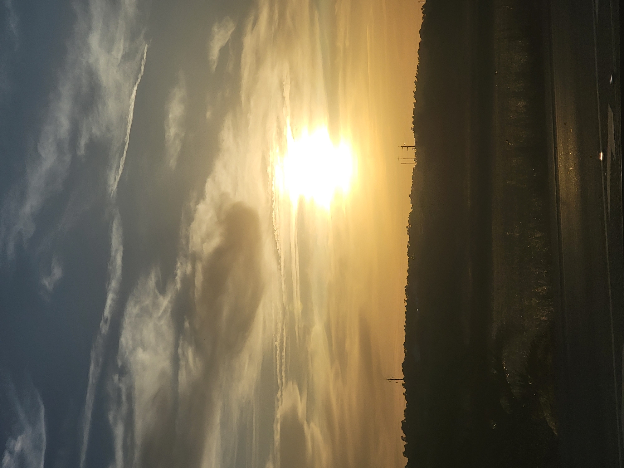 The image captures a sunset over a landscape that appears to be on the outskirts of Faro, Portugal. The sky is dominated by a bright, radiant sun positioned in the upper center of the frame, surrounded by a dramatic display of clouds. The clouds vary in color from deep blues and grays in the upper left to soft yellows and oranges closer to the sun, creating a gradient effect. Some clouds are wispy and elongated, while others are more voluminous and textured.
Below the sky, a dark, silhouetted hill or ridge stretches across the middle ground, topped with a line of trees and vegetation. Perched on the crest of this ridge are two tall, slender structures, likely electricity pylons or transmission towers, which are also silhouetted against the bright sky.
The foreground depicts a dark, shadowy area, possibly a field or open ground, with faint glints of light scattered across its surface. The very bottom of the image shows a dark, reflective surface, which could be a road or a body of water, with a few bright streaks of light suggesting illumination or reflections. The overall mood is serene and evocative, typical of a sunset scene. There are no people or animals visible in the frame.