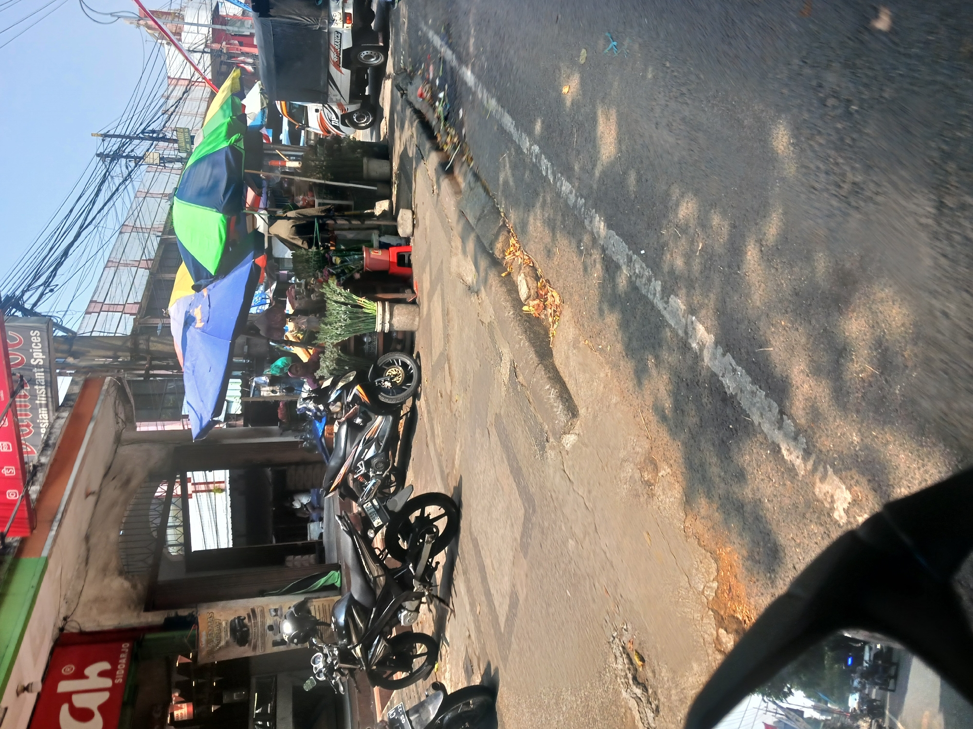 The image depicts a street scene in Singosari, Indonesia, likely a commercial area. In the foreground, the reflection in a rearview mirror shows a street with several motorcycles and a white truck. The primary focus of the image, however, is the street on the right side of the road. Several motorcycles are parked on a raised concrete sidewalk in front of a building with awnings. Two large, colorful umbrellas are set up, one predominantly blue and the other green and yellow, casting shade over stalls that appear to be selling plants and other goods. A white truck with "PERFORMANCE" printed on its side is visible in the background, parked on the street. The street itself is paved with asphalt and shows signs of wear, with debris and fallen leaves scattered along the curb. Electrical wires are strung overhead, common in urban Indonesian settings. A sign above the building reads "Instant Spices" and another sign in red letters appears to say "Ah" with "SIDOARJO" underneath. The overall impression is a busy, sunny day in an Indonesian town, with commerce happening on the sidewalks and traffic moving on the road.