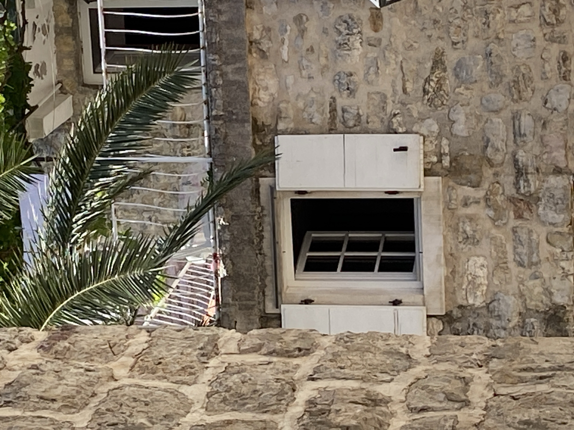 This image, captured in the city of Budva, Montenegro, presents a detailed view of traditional stone architecture under bright daylight. The foreground is dominated by a rough-hewn, irregular stone wall on the left side, displaying varying shades of beige and gray. To the right of this wall, a cream-colored wooden window with a multi-pane casement is set into a second wall of irregular stone masonry. The left side of the casement is partially open, revealing a dark interior, while both white wooden shutters are fully open, with the right shutter resting flat against the wall. Dark, simple hinges are visible on the shutters. A large, arching green palm frond descends from the upper left, diagonally traversing the mid-section of the frame and partially obscuring a white metal balcony railing in the upper right background. Behind the railing, a dark rectangular opening, indicative of a doorway or window, is visible within another stone wall, topped by a flat concrete or stone slab. Additional green leafy foliage is present above this upper structure. The scene is devoid of people, animals, or vehicles.