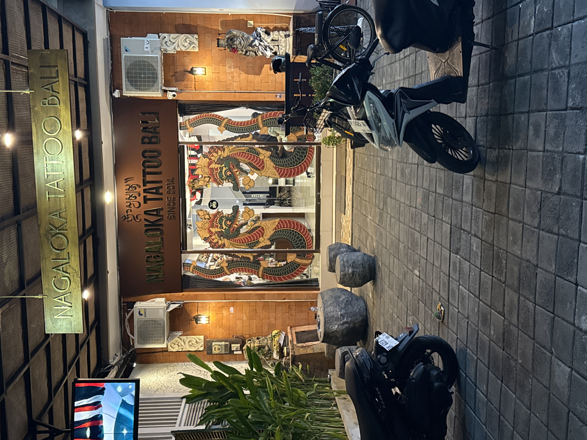 The image depicts the exterior of "Nagaloka Tattoo Bali," a tattoo parlor located in Denpasar, Indonesia. The shop's name is prominently displayed in gold lettering on a wooden sign above the entrance and also on the glass storefront. The storefront features large glass doors adorned with intricate artwork of what appear to be Naga serpents, a common motif in Balinese art.
In front of the shop, several motorcycles are parked on a tiled walkway. One black motorcycle is parked to the left, and a lighter-colored scooter is parked to the right. A black bicycle is also visible to the right of the scooter. A round stone table and three cylindrical stone seats are situated in front of the entrance, suggesting an outdoor seating area.
The shop's facade is constructed of red brick. Above the glass doors, two air conditioning units are mounted on the brick wall. To the left of the entrance, a large potted plant with tall green leaves stands next to a decorative stone pillar. Various other decorative elements, including statues and lanterns, are visible around the entrance and on the brick wall.
The lighting suggests it is evening or night, with artificial lights illuminating the storefront and the surrounding area. The overall scene conveys a sense of a typical commercial establishment in a tropical urban environment.