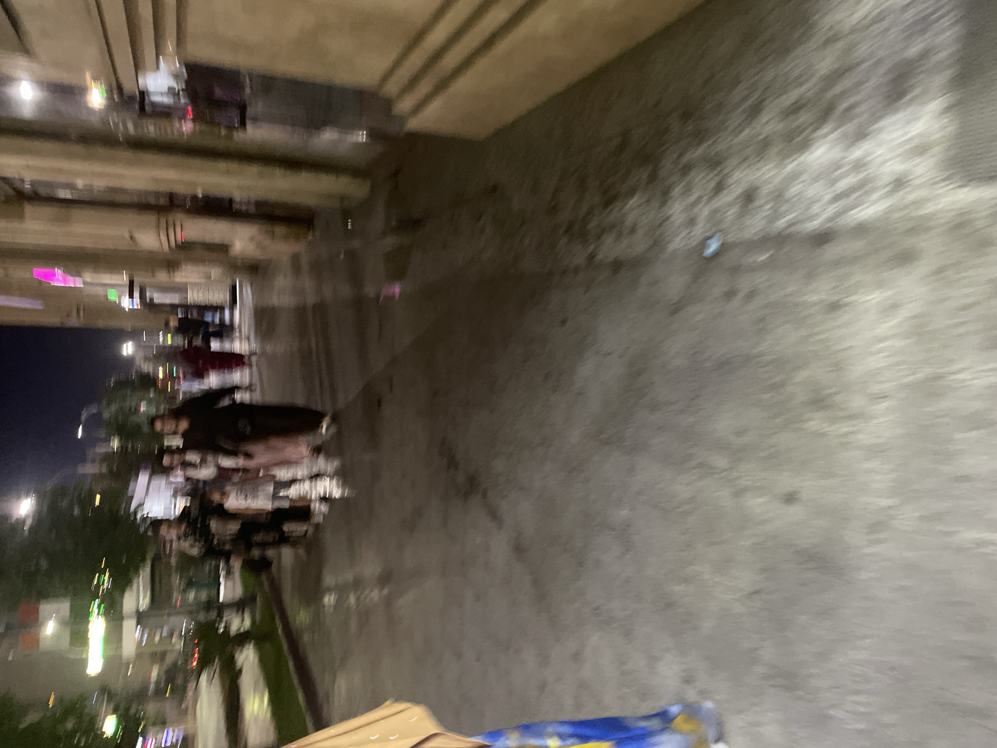 The image depicts a nighttime scene on a city street in Dushanbe, Tajikistan, likely in an area with commercial buildings. A group of people, including adults and a child, are walking on a wet sidewalk. The lighting suggests it has recently rained or is damp. The people are dressed in casual attire. Some buildings are visible along the right side of the street, with lights indicating businesses are open. The overall atmosphere is somewhat blurred, suggesting movement or low light conditions.