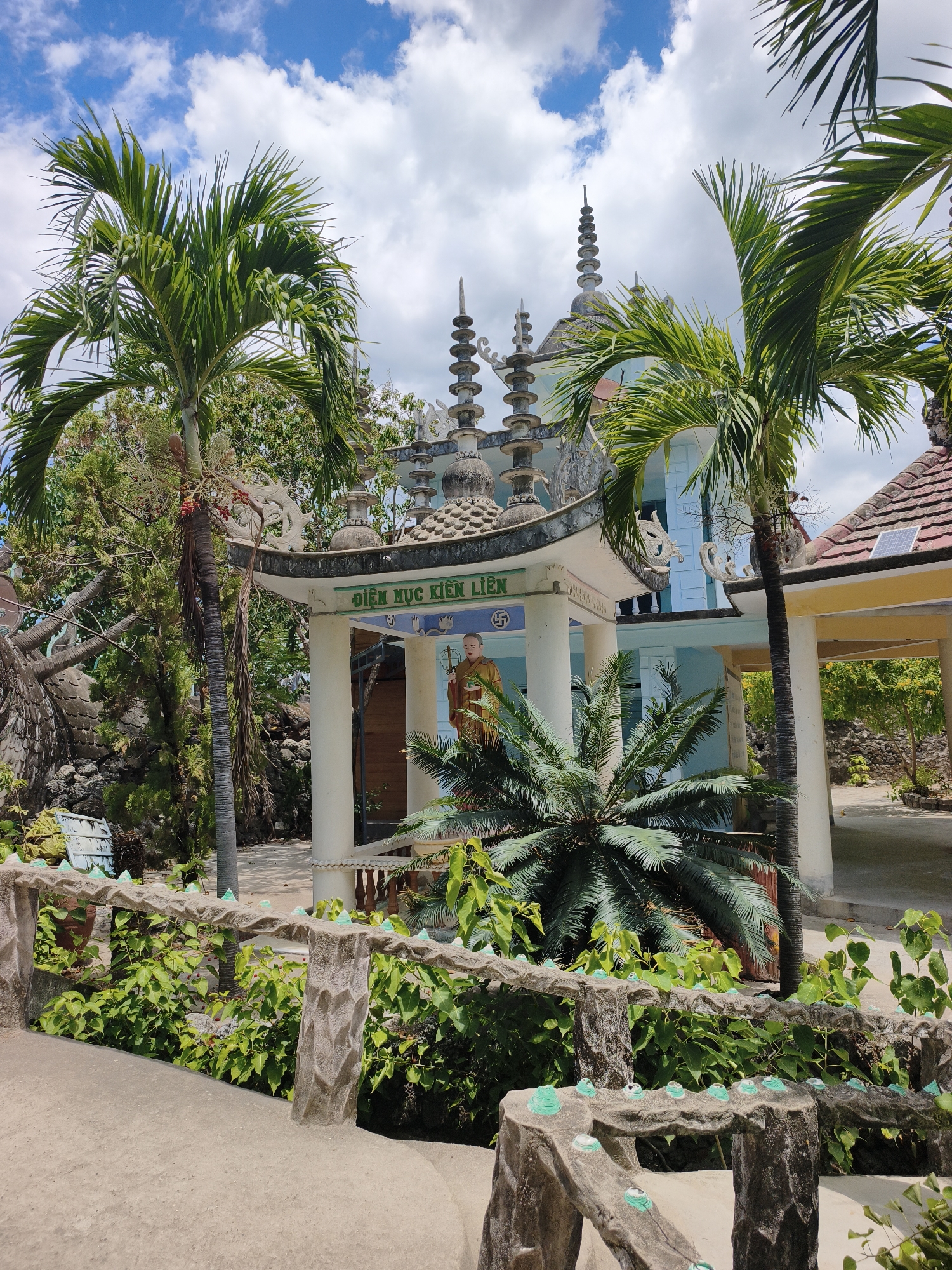 The image depicts an outdoor scene in Cam Ranh, Vietnam, featuring a temple-like structure with distinct architectural elements. The building, painted in light blue and yellow, is adorned with multiple tiered roofs and decorative spires. A prominent sign with Vietnamese characters is visible above an open-air pavilion. Within this pavilion, a statue of a robed figure, likely a monk, stands. The foreground is occupied by a low, textured concrete barrier decorated with small, green, rounded objects, bordering lush green foliage. Several palm trees, some with fronds obscuring the sky, are scattered throughout the scene. The sky above is a mix of blue and white clouds. The overall environment suggests a warm, sunny day.