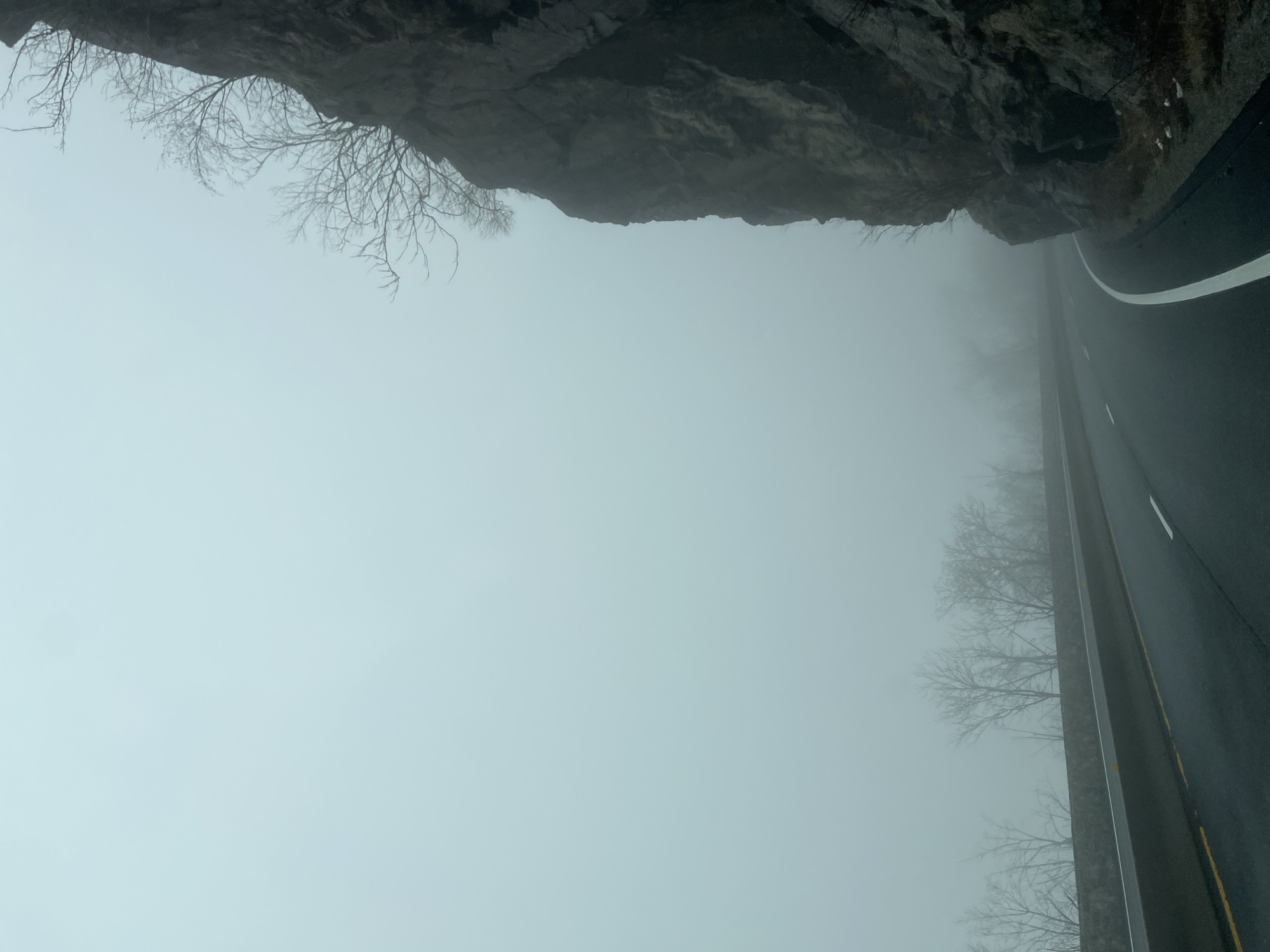 The image shows a road in West Point, United States, shrouded in dense fog. On one side of the road, a rocky cliff overhangs the asphalt, with bare tree branches silhouetted against the sky. The road is wet and marked with white and yellow lines. The fog obscures the surrounding landscape, limiting visibility to a short distance. The scene suggests a remote or mountainous location.