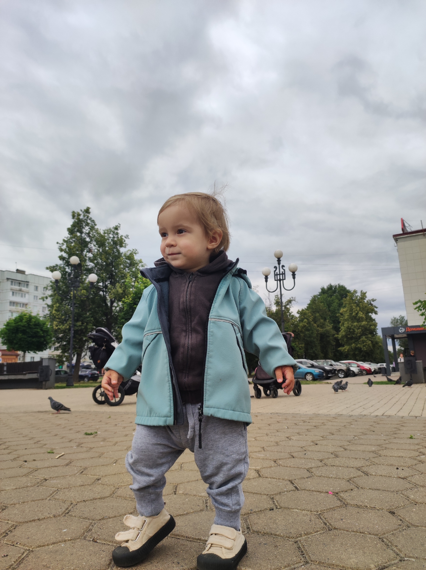 A low-angle, full shot captures a toddler standing on a paved square in Chaikovsky, Russia, under a cloudy sky. The toddler, facing right, wears a light blue hooded jacket over a dark hooded sweatshirt and gray sweatpants. Their feet are clad in cream-colored shoes with Velcro straps and black soles. To the left of the toddler, a dark stroller is partially visible. In the background, several cars are parked, and trees with green foliage line the area. A building with white siding and a red sign is to the right. Pigeons are scattered across the square in the foreground and midground.