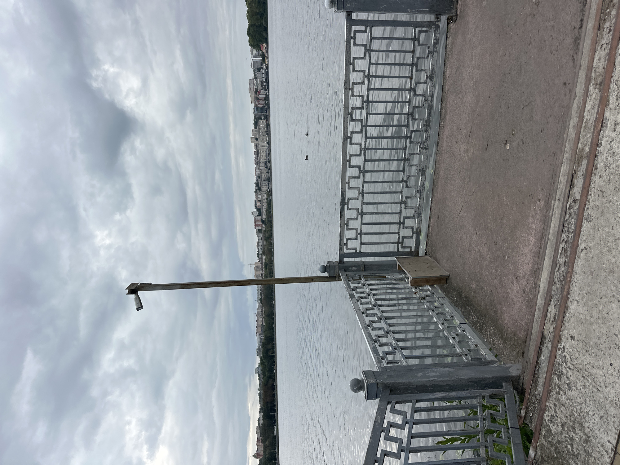 The image depicts a waterfront view of Ternopil Lake, also known as Ternopil Pond, in Ternopil, Ukraine. In the foreground, there is a concrete walkway with a decorative metal railing, a common feature along the lake's embankment. The railing is gray and features a geometric pattern. A utilitarian wooden board is propped against the railing. A metal pole, likely for lighting or surveillance, rises from the walkway.
The lake occupies the midground, its surface rippled by a light breeze. Two ducks are visible swimming on the water. In the distance, the cityscape of Ternopil stretches along the opposite shore. Buildings of varying heights and architectural styles form a dense urban skyline, punctuated by trees and greenery. The sky is overcast with heavy, gray clouds, suggesting a cool or potentially rainy day. The overall impression is one of a calm, urban waterfront on a cloudy day.