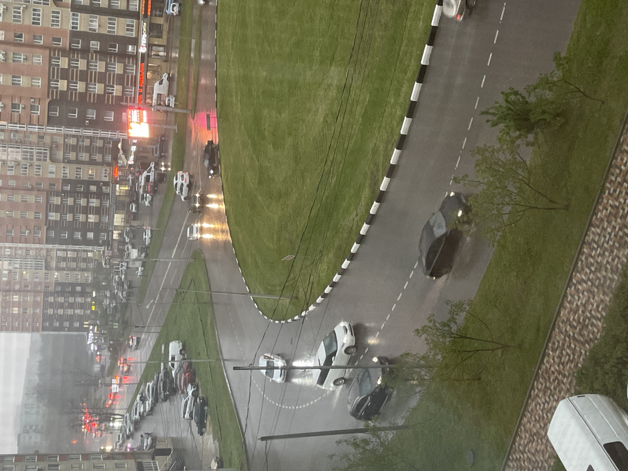 The image depicts a rainy street scene in Stavropol, Russia, viewed from an elevated perspective. The street is wet and reflections of car headlights are visible. Several cars are moving along the road, while others are parked on the side. There are apartment buildings visible in the background, with signage and red lights illuminated. Green spaces, including grassy areas and trees, border the road. A curb with black and white stripes lines a portion of the road. Overhead wires are also visible. The weather is overcast and visibility is reduced due to the rain.