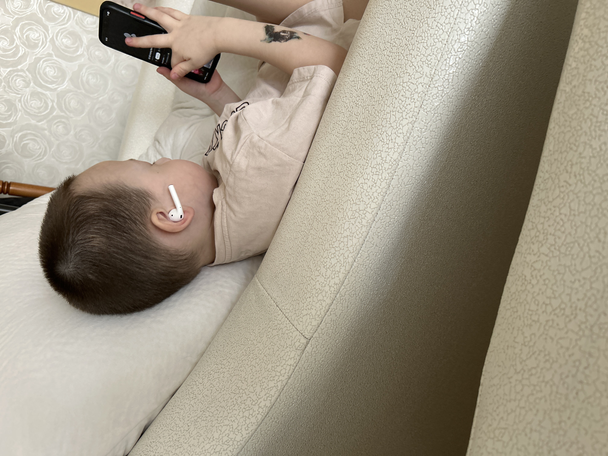 An overhead-angle image depicts a young child, approximately two to four years old, reclining in an indoor setting. The child, with short brown hair, is wearing a light-colored short-sleeved t-shirt and has a dark, temporary tattoo of an animal's head (possibly a dog or wolf) on the left bicep. A white wireless earbud is inserted into the child's right ear. The child holds a black smartphone with both hands, positioned over the screen. The phone displays a media player interface with a white central icon and text in a non-English script, along with a "19:29" timestamp in the upper left corner of the screen. The child's head rests on a plain white pillow or cushion, positioned on a light beige or off-white piece of furniture with a textured, mottled surface, likely a sofa or armchair. In the background, a wall covered with light-colored, patterned wallpaper featuring a repeating floral or rose design is visible. A dark wooden object, possibly furniture, is partially visible in the upper right. The scene's specific location in the city of Chaikovsky, Russia, cannot be determined from the visual information presented in the image.
