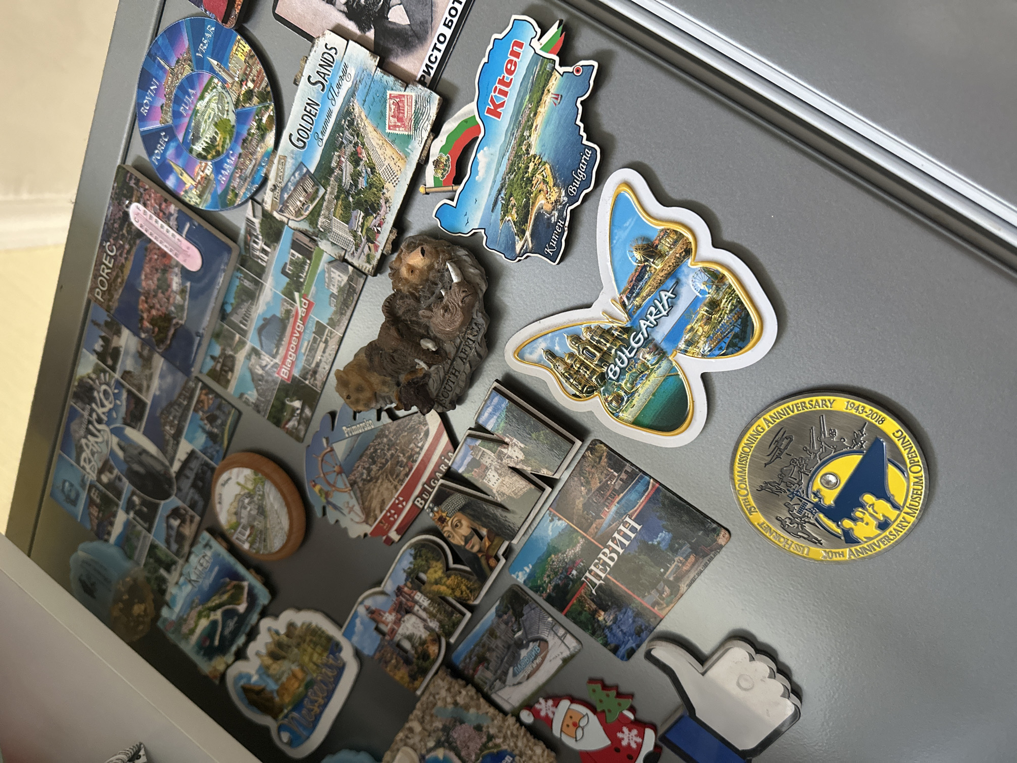 A gray refrigerator door is covered in various souvenir magnets. The magnets feature locations such as Poreč, Rovinj, Vrsar, Pula, Bangkok, Kiten, Blagoevgrad, Primorsko, Nessebar, Golden Sands, and Devin. There are also magnets related to Bulgaria, including one shaped like a butterfly and another shaped like the country's outline. A coin-shaped magnet commemorates the USS Hornet. Other magnets include a stone lion, a Santa Claus, and a Facebook "thumbs up" icon. The location in Marchaevo, Bulgaria, is not indicated or depicted in the image.
