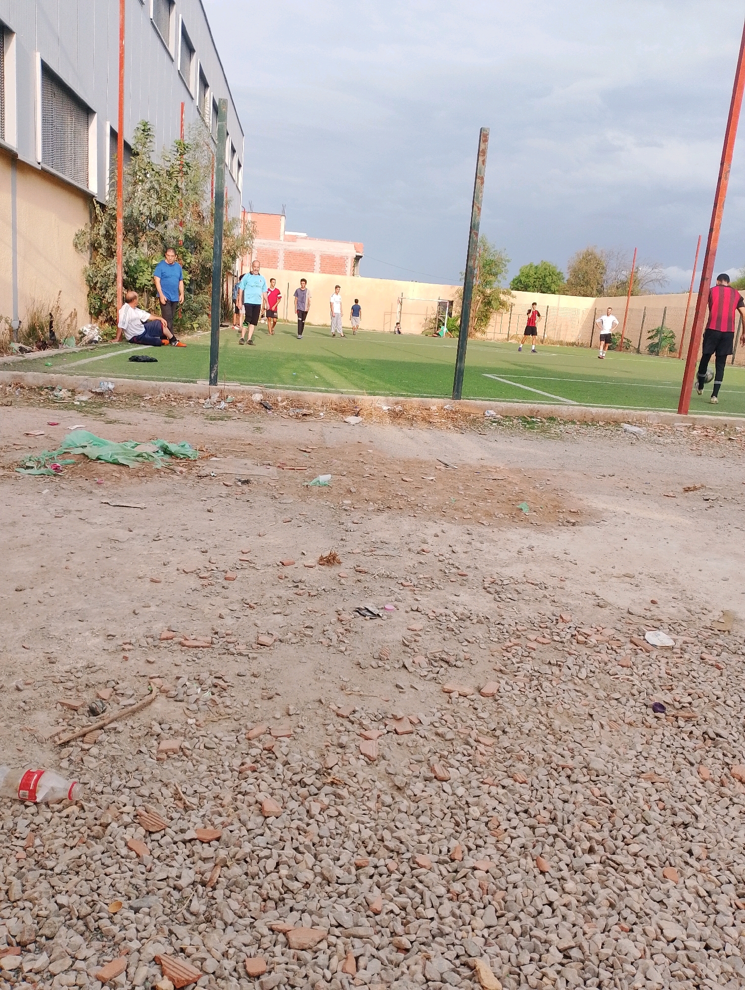The image depicts an outdoor artificial turf soccer field adjacent to a building with a gray, corrugated metal exterior and a beige lower wall. The field is bordered by a chain-link fence. Several individuals are present, some actively engaged in soccer-related activities and others observing.
On the field, there are at least seven individuals visible. Two are in the foreground, one wearing a red and black striped shirt and black shorts, and the other in a white shirt and black shorts. Further back, several more individuals are scattered across the field. One person in a blue shirt appears to be sitting on the ground near the edge of the field, next to a person in a white shirt and blue shorts. Another individual in a blue shirt is standing on the field.
The environment surrounding the field is a mixture of rough terrain with gravel and broken bricks, littered with debris including a plastic bottle with a red label. Green netting or plastic sheeting is also visible on the ground. The sky is overcast with clouds. The scene appears to be a public or community sports area in Bordj Menaiel, Algeria.