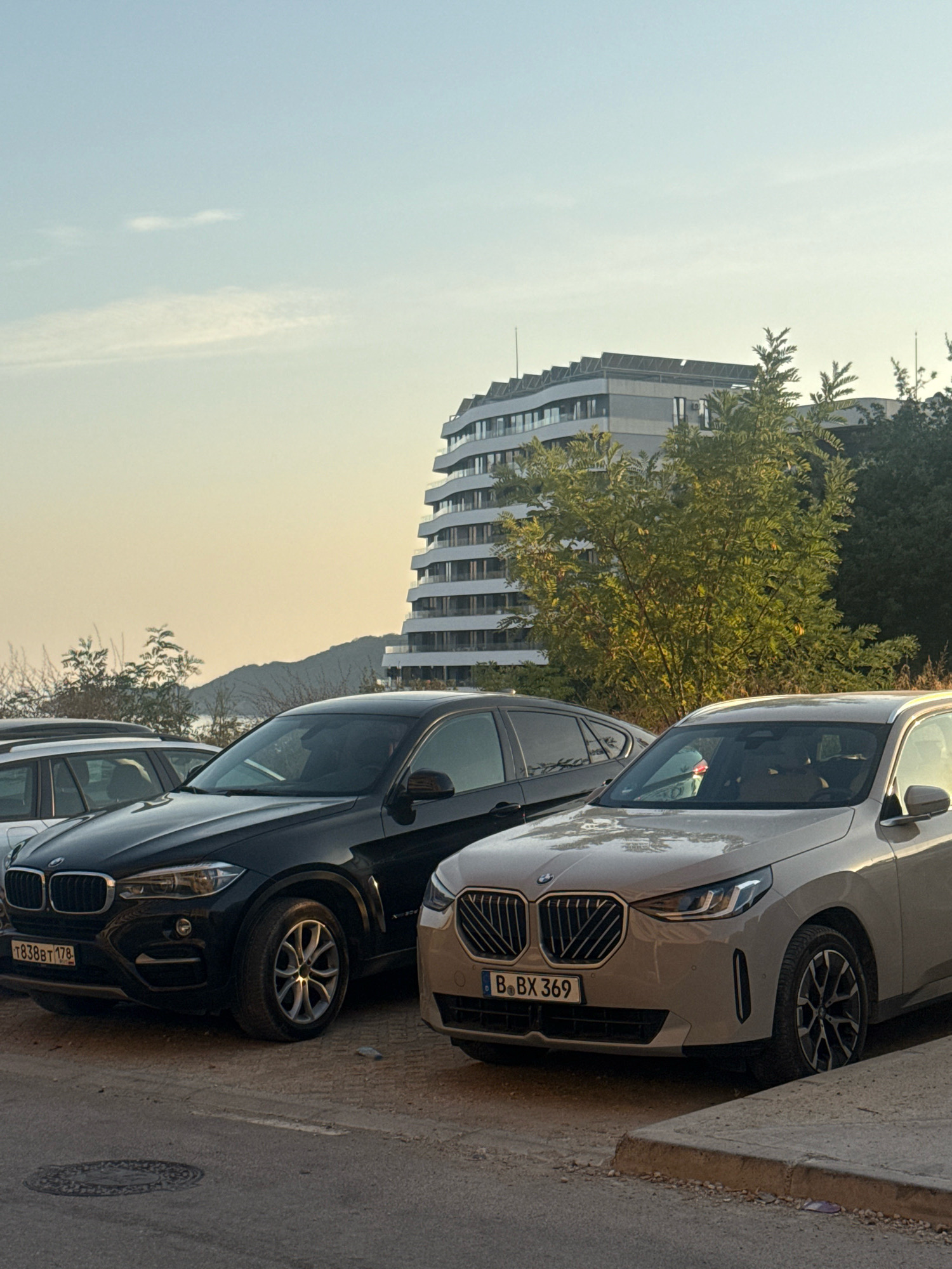 The image depicts an outdoor scene, likely in the city of Boreti, Montenegro. In the foreground, two BMW cars are parked on a gravelly surface adjacent to a paved road with a curb. The car on the left is black, and the car on the right is a light tan color. The black car's license plate reads "T838BT 178". The tan car's license plate reads "B. BX 369". Behind the cars, a multi-story modern building with a distinctive curved facade is visible, surrounded by lush green trees. In the background, hills are visible against a pale sky with some wispy clouds. The lighting suggests it is either early morning or late afternoon, with a warm, soft glow illuminating the scene. No people are clearly visible within the cars or the immediate vicinity. The overall impression is one of a residential or commercial area with a backdrop of natural landscape.