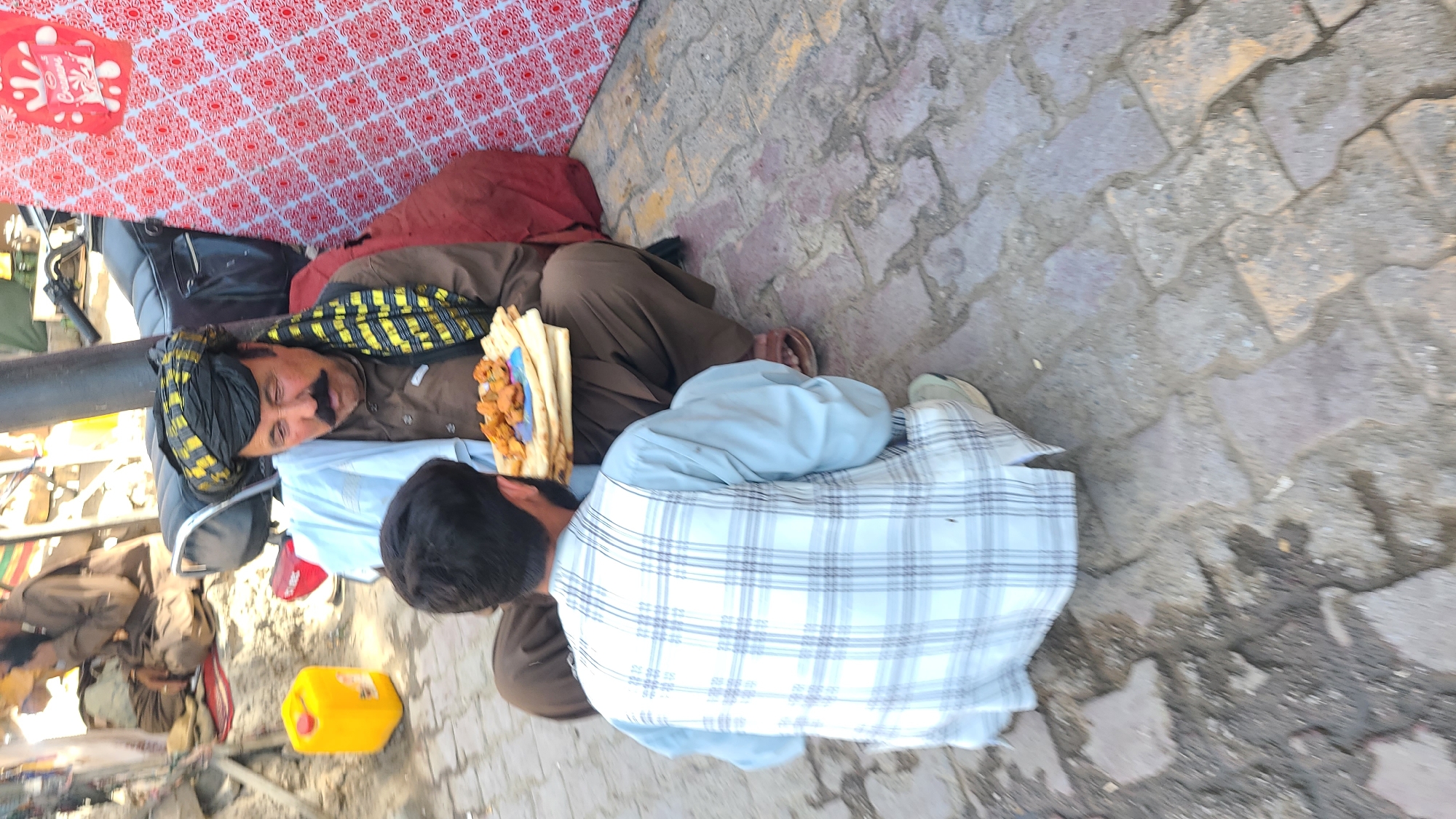 A man wearing a brown shalwar kameez and a black and yellow turban lies reclining against a black motorcycle seat. He holds a piece of naan bread topped with fried food. Another man wearing a light blue and white plaid shirt sits beside him. They are on a paved street in what appears to be the city of Quetta City Tehsil, Pakistan. A yellow gas can and other debris are nearby. The background shows a red and white patterned cloth and possibly a building or structure.
