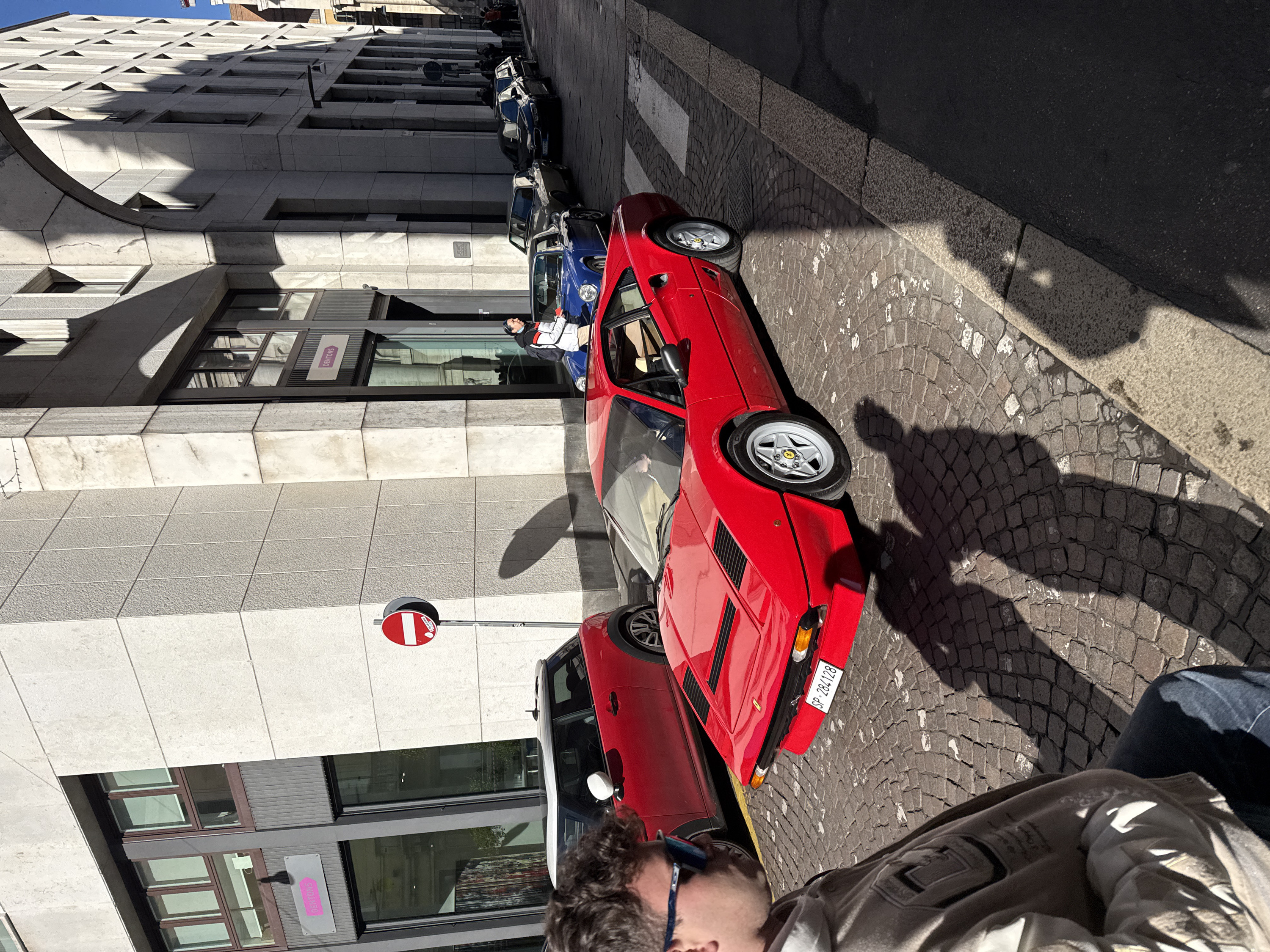 The image displays a sunlit street scene in Milan, Italy, featuring cobbled pavement and multi-story stone-clad buildings under a clear blue sky.
In the immediate foreground on the left, a male subject is partially visible, wearing blue mirrored sunglasses, a light-colored hooded sweatshirt, and dark jeans, looking towards the street. Prominently positioned in the midground is a red Ferrari 308 GTB/GTS, identifiable by its distinctive bodywork and an Italian license plate "SP 284128" affixed to its front. The vehicle is equipped with silver five-spoke alloy wheels, and a driver is visible within the cabin.
To the left of the Ferrari, a red Mini Cooper with a white roof is parked. To the right of the Ferrari, a pedestrian walks on the cobblestones, dressed in a white jacket with black and red accents, dark pants, and a light blue face mask, carrying a dark backpack. Further down the street, a blue Porsche 911 is parked, followed by a line of other vehicles, including a dark classic coupe and a dark BMW sedan bearing the Italian license plate "ET 211SH". Additional pedestrians are visible further along the street on the right.
A red circular "no entry" sign with a white horizontal bar is mounted on a pole to the left of the Ferrari. The buildings lining the street feature light-colored stone or concrete facades with rectangular, dark-framed windows. A visible sign for "DENTONS" with a pink arrow is affixed to a building on the left. On the right side, an architectural archway spans overhead, casting sharp shadows across the facade and street. A sign for "ASSICURAZIONI GENERALI" is partially visible on a building further down the street on the right. Strong sunlight from the upper left casts distinct, elongated shadows across the pavement and building surfaces.