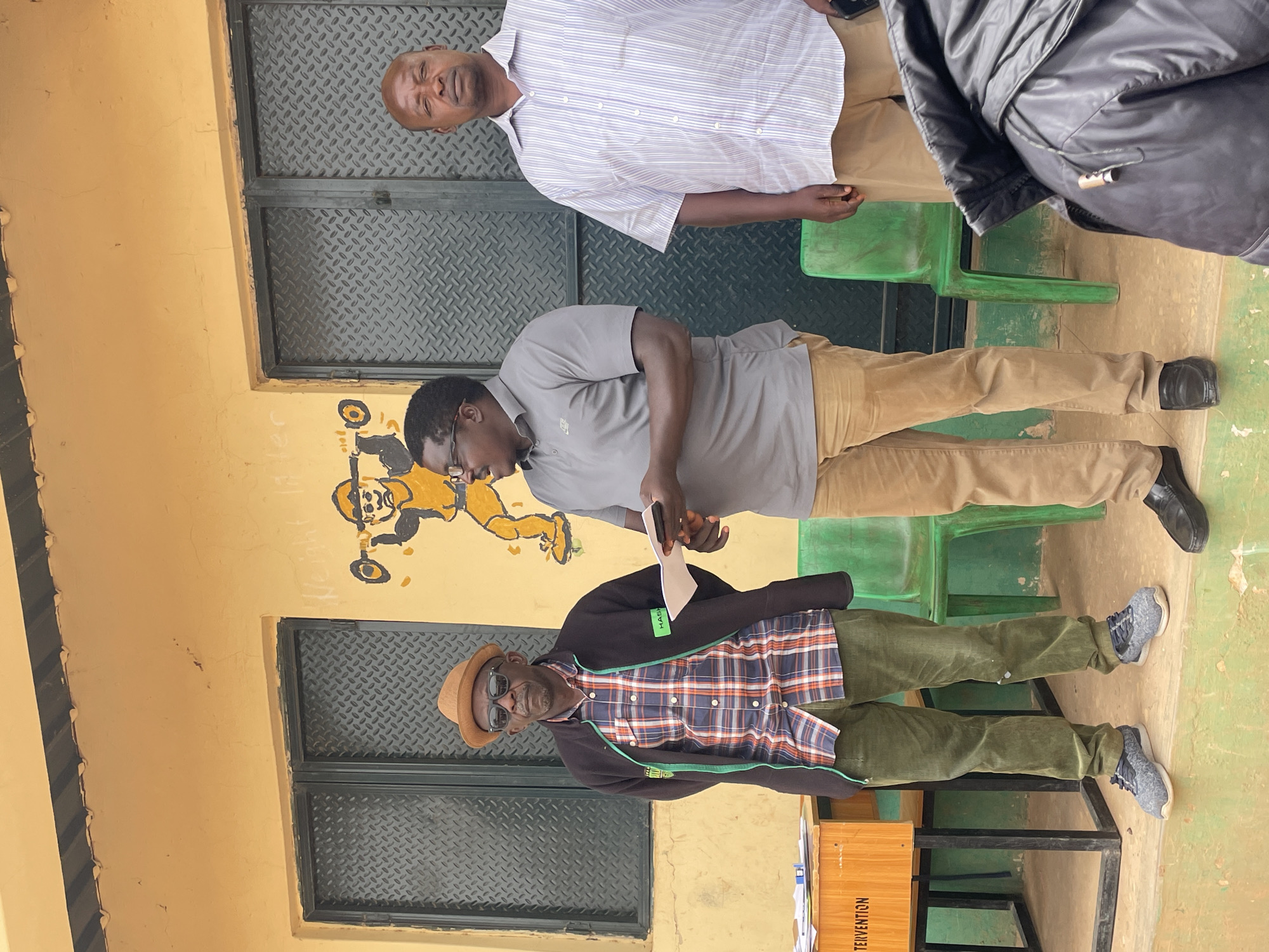 Three men are standing outdoors against a yellow building with dark gray metal doors or windows and a painting on the wall. The man closest to the camera wears a hat, sunglasses, a plaid shirt under a dark jacket with a green trim, and green pants. He stands next to a wooden box with writing on it. The second man is wearing a gray polo shirt and tan pants, holding a white object in his hands. The third man is wearing a light-colored striped shirt and tan pants. The background is blurry and unclear.
The location is Unknown, Unknown.