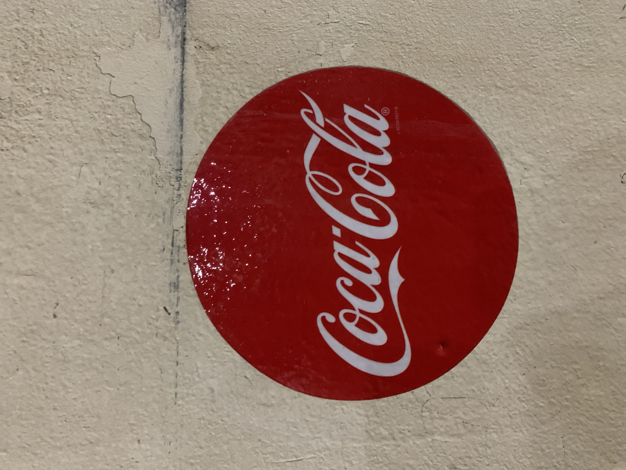 The image displays a glossy, circular red Coca-Cola sign mounted on a textured, light-colored wall in Santa Ana, Venezuela. The sign features the iconic "Coca-Cola" logo in white script, oriented vertically. A small registered trademark symbol (®) and the alphanumeric code "J-2038362-6" are visible in white text near the top right of the logo. The red surface of the sign is reflective, showing glares and an uneven, possibly wet or heavily coated, texture. A small indentation, likely a screw or nail hole, is present near the bottom right edge of the sign, indicating its attachment.
The surrounding wall is off-white or beige, characterized by a rough, weathered texture with visible cracks, flaking paint, and general signs of age and wear. A prominent dark vertical line or groove runs down the wall to the left of the sign, possibly a structural seam or a previous marking. The setting appears to be a static outdoor or semi-outdoor location, given the condition of the wall. No people or specific activities are depicted; the focus is entirely on the Coca-Cola signage and its immediate, worn environment.