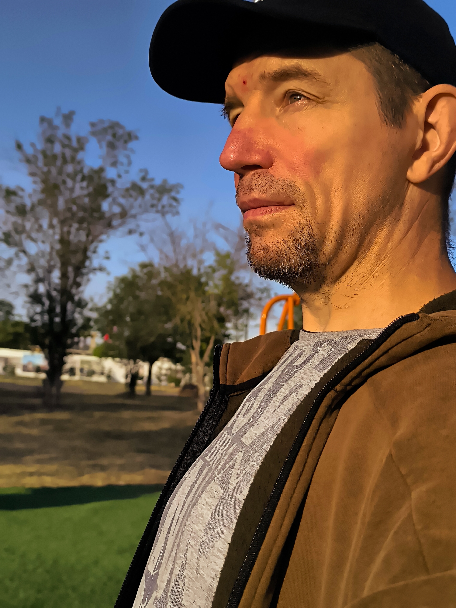 A close-up, profile shot features a man wearing a black baseball cap and a brown hooded jacket. The man's face is illuminated by warm, golden light, highlighting his features and a small red mark on his forehead. He has stubble on his chin and a beard. In the background, out-of-focus trees and a blue sky suggest an outdoor setting. The overall scene captures a moment of the man looking off to the side.