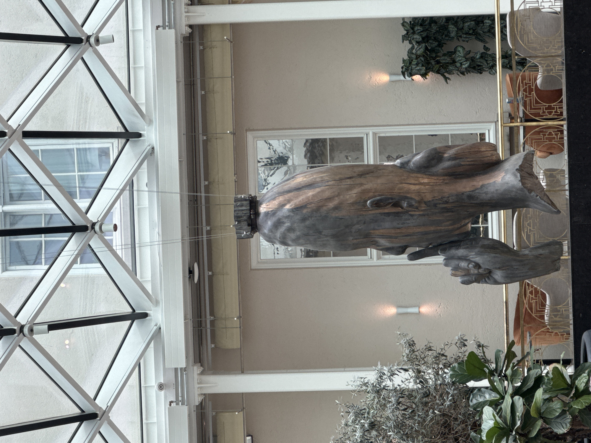 The image shows a large, abstract sculpture of a head in profile with a finger to its lips, in a gesture of silence. The sculpture is made of a dark, metallic material with hints of wood grain, and is suspended from the ceiling. The background features a modern, glass-paneled atrium with a white, geometric ceiling structure. Two windows with multi-paned glass are visible on the neutral-colored wall behind the sculpture. The scene is illuminated by ambient light filtering through the glass ceiling and by two vertical wall sconces, one on either side of the windows. In the foreground, there are lush green plants and an art deco-style railing. The sculpture appears to be located in a public indoor space, possibly a hotel lobby or museum in Copenhagen, Denmark.