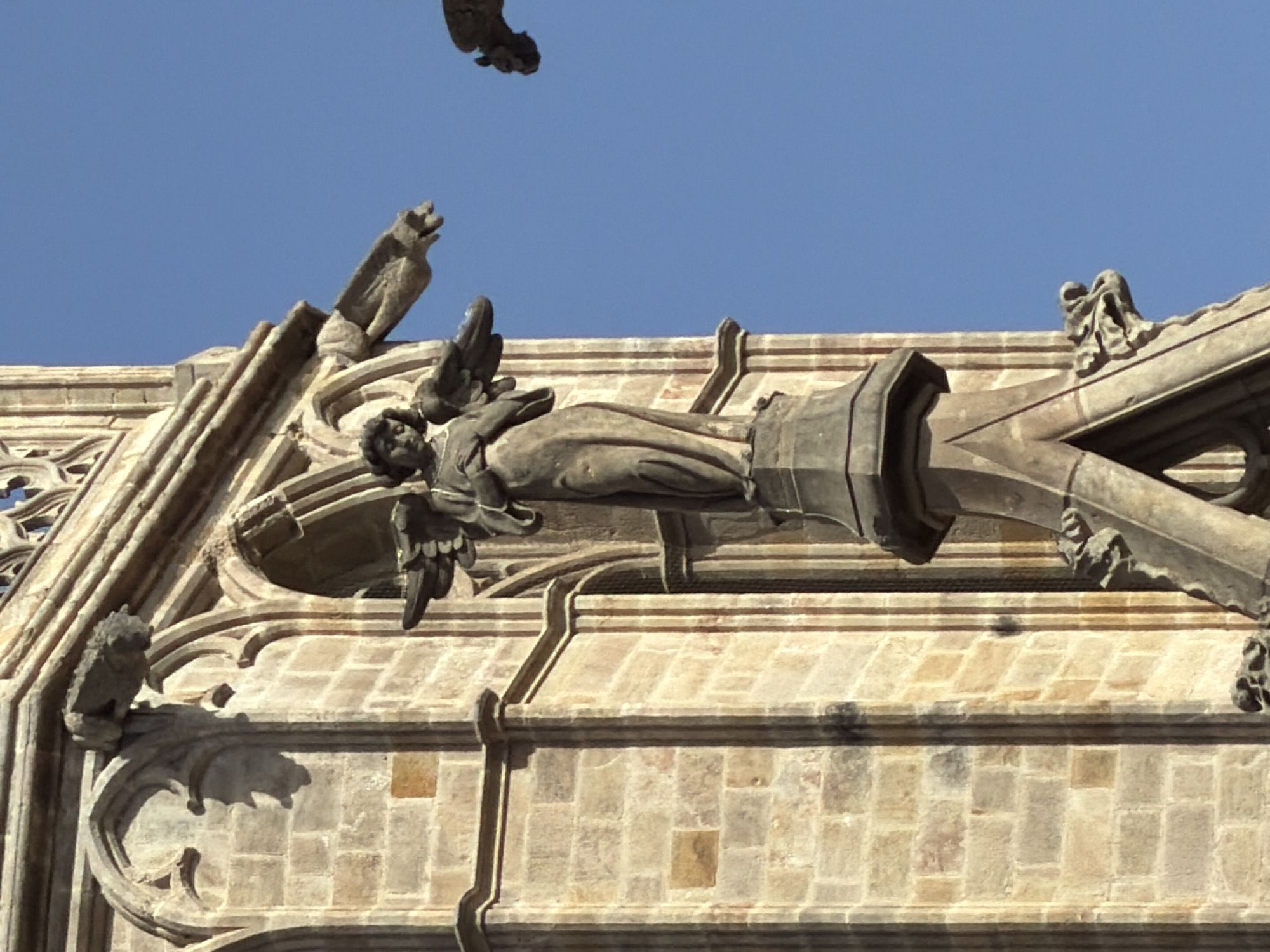 This is a low-angle, outdoor, medium shot of a statue of an angel on a tall, stone building, possibly a cathedral, against a clear blue sky. The angel is made of stone and is depicted with wings and draped clothing, with its hands held in front of its chest. It is positioned on a decorative stone pedestal. The building is constructed of light-colored stone bricks and features Gothic architectural elements, including arched windows and ornate carvings. Gargoyles are visible protruding from the building's facade on either side of the angel statue. The overall lighting suggests it is daytime. The building is likely part of Barcelona Cathedral, given the architectural style and presence of such decorative elements.