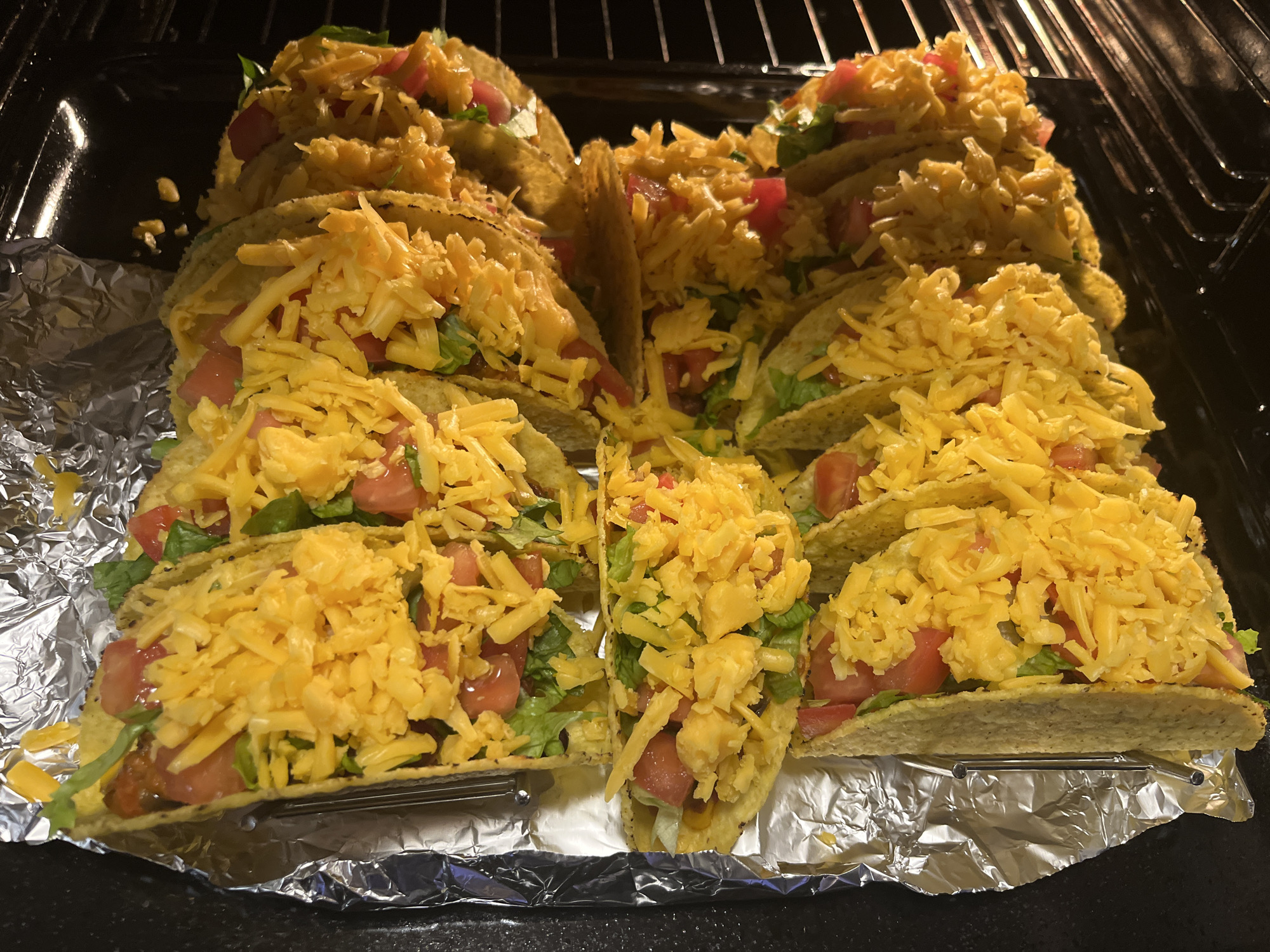 This media file, originating from Veliko Tarnovo, Bulgaria, captures a close-up, high-angle view of numerous assembled hard-shell tacos, likely in the process of being heated in an oven. The tacos are neatly arranged side-by-side in a silver metal taco holder, which rests on a sheet of crumpled aluminum foil.
Each taco is generously filled with a vibrant mix of ingredients. The most prominent topping is a thick layer of shredded yellow cheese, sprinkled over diced red tomatoes and shredded green lettuce. A darker filling, possibly ground meat, can be glimpsed beneath these toppings in several of the tacos.
In the background, dark metal oven racks are visible, confirming the setting as an oven or toaster oven. The aluminum foil beneath the taco holder suggests preparation for baking or to catch any drippings. The lighting is bright, highlighting the texture of the cheese and the fresh colors of the vegetables, while the background remains dark, focusing attention on the food. No visible text is present in the image.