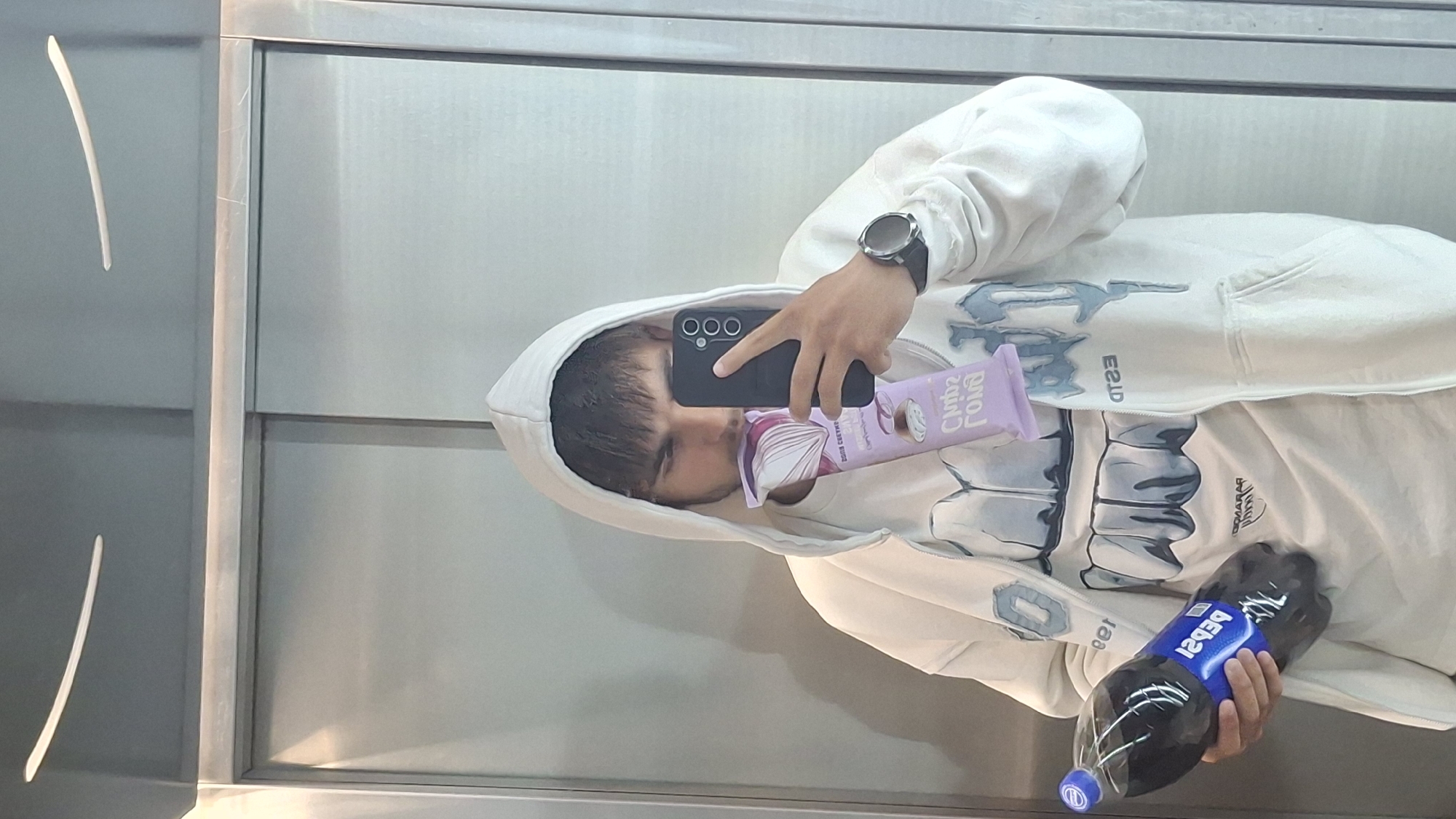 A person wearing a white hoodie and a watch is taking a selfie in what appears to be an elevator. They are holding a purple snack bar in one hand and a large bottle of Pepsi in the other. The selfie is being taken with a black smartphone. The elevator interior consists of metal panels. There is no information in the image to confirm the location is Varna, Bulgaria.