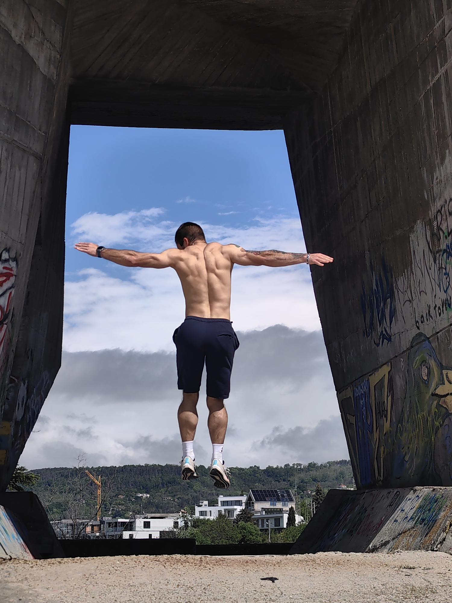 A man is suspended in mid-air, arms outstretched, seen from the back. He is wearing dark shorts, white socks, and athletic shoes. He appears to be within a large concrete structure with graffiti on the walls, framing a view of a cloudy sky and a landscape with buildings and trees. Based on the context of Varna, Bulgaria, and visible landmarks, the location is likely the Asparuhov Bridge area.