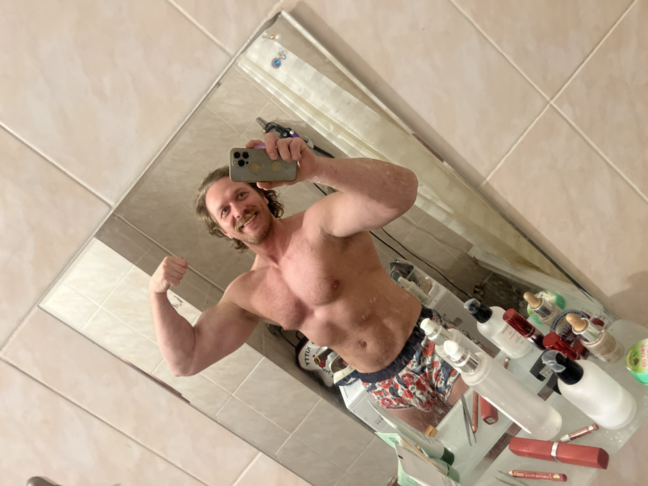 A muscular, shirtless man poses in front of a bathroom mirror, flexing his left bicep. He is holding a smartphone, presumably taking the picture. He sports a mustache and short, wavy hair. His colorful, patterned shorts are visible. The bathroom counter is cluttered with various skincare products. The mirror reflects the bathroom tiles and a shower curtain.