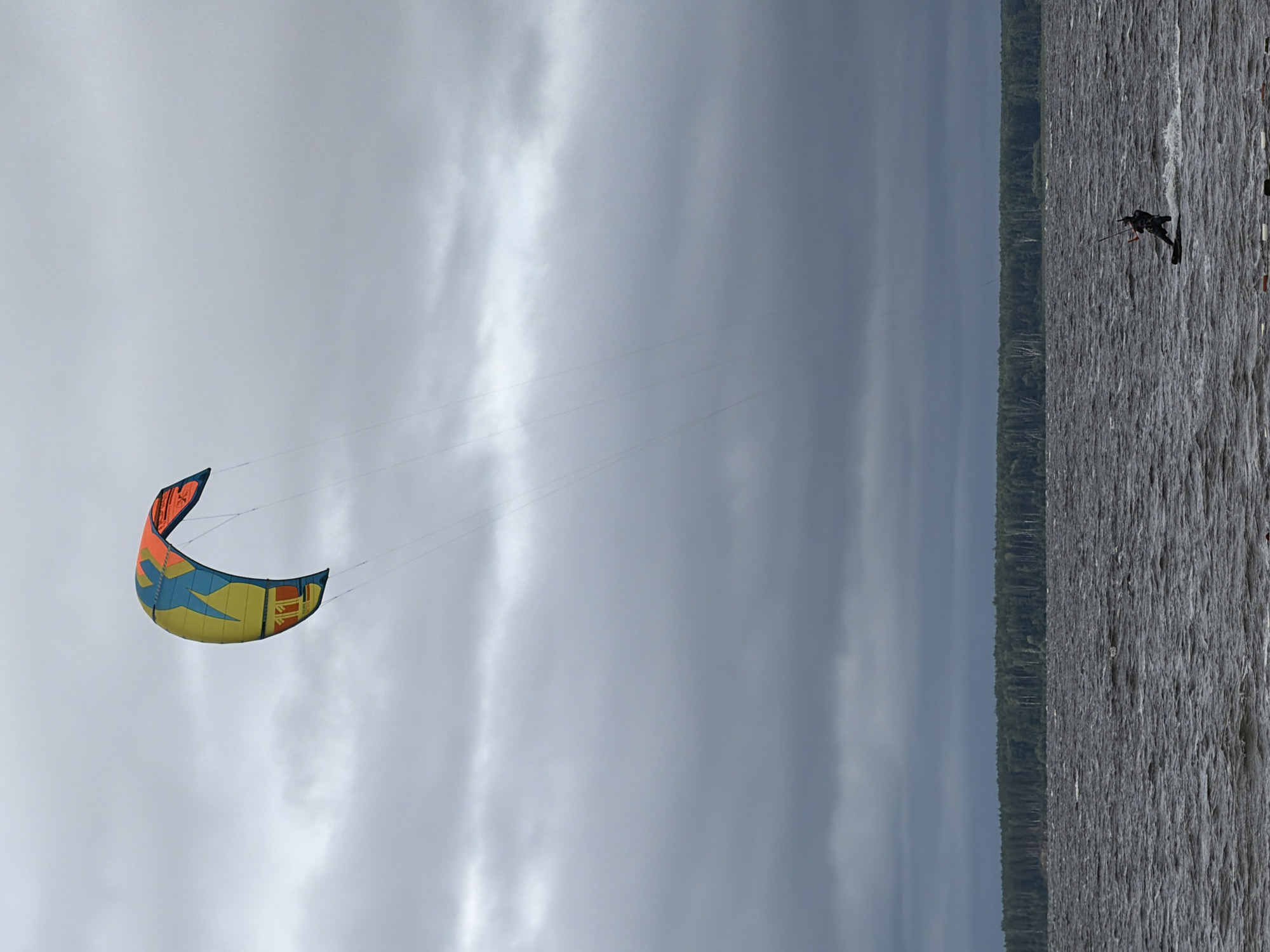 A single kiteboarder navigates choppy waters on a cloudy day. The kiteboarder, wearing dark clothing, is positioned in the lower right quadrant of the frame, leaning back as they ride across the surface of the water. The kite, a brightly colored structure of yellow, blue, and orange, is suspended high in the sky, tethered to the kiteboarder by thin lines. Behind the water, a dense line of dark green trees forms a stark horizon against the overcast sky. The overall impression is one of wind-powered activity against a dramatic, atmospheric backdrop, suggestive of a location like the Kama River in Chaikovsky, Russia, known for its watersports.