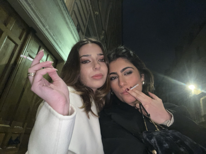 Two young women stand close together outside at night, each holding a lit cigarette. The woman on the left wears a white coat and has light brown hair. The woman on the right wears a black coat and has dark brown hair, and a handbag is visible. A building with closed doors is partially visible on the left side of the image, and street lights illuminate the background.