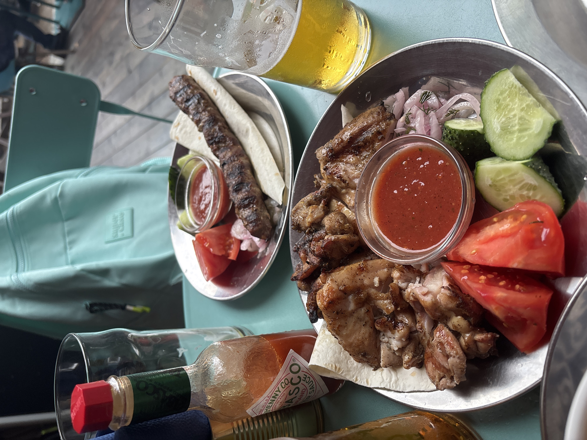 The image depicts a close-up view of a table setting with food and drinks, likely at an outdoor cafe in Komarovo, Russia. In the foreground, a metal plate holds a serving of grilled chicken pieces, sliced tomatoes, cucumber slices, red onions, and a small bowl of red sauce. Another metal plate in the background contains a grilled meat skewer, flatbread, and a similar red sauce. A bottle of Tabasco sauce is visible to the left of the foreground plate. Two glasses of beer, one partially full and the other with condensation, are also present. Behind the food, a light blue backpack rests against a chair. The table appears to be teal, and the flooring, visible in the background, is wooden planks. The overall scene suggests a casual meal outdoors.