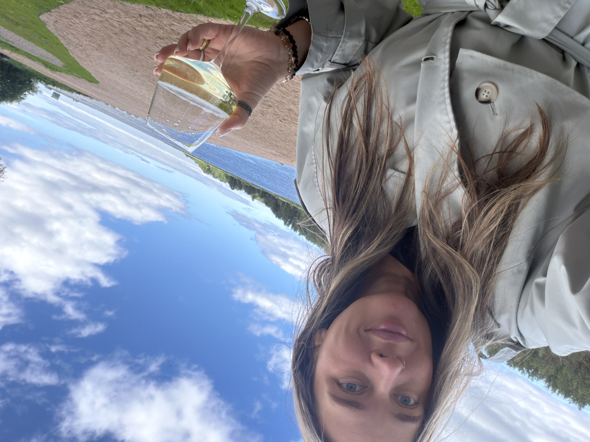 A woman is taking a selfie outdoors, holding a glass of white wine. She is wearing a light-colored trench coat and has long, blonde hair. The background shows a beach, a body of water, and a treeline under a cloudy blue sky. The location is identified as Solov'evo, Russia.
