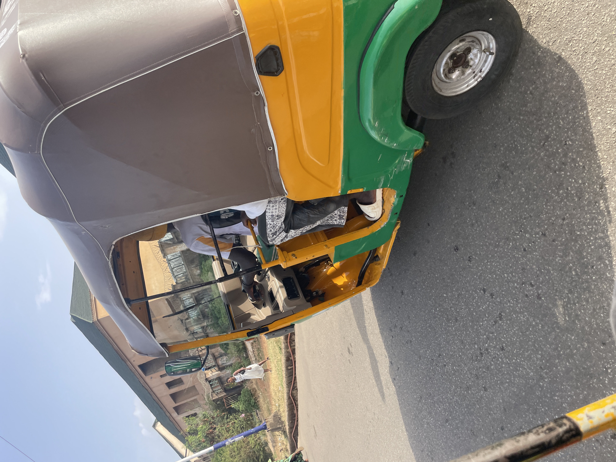The image shows a three-wheeled vehicle, possibly a Bajaj RE, on a paved road. It has a brown roof, yellow sides, and green fenders. A passenger is visible inside the vehicle, wearing a white shirt and black pants. The driver is visible at the steering wheel. Outside the vehicle, a person in a white dress walks on the roadside near a building. The scene appears to be in Jos, Nigeria. Vegetation and a blue and white striped pole are visible in the background, suggesting a city environment. The pavement is asphalt.