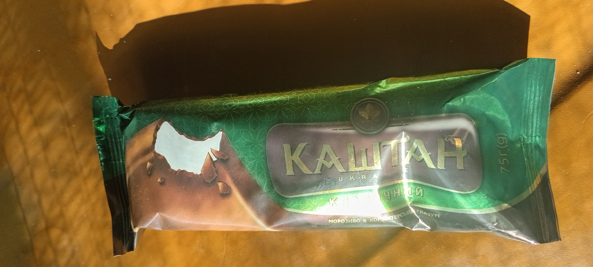 The image shows a close-up, top-down view of a chocolate ice cream bar wrapper on a brown, textured surface, possibly wood, illuminated by a strong light source casting a dark shadow to the right. The wrapper is predominantly green with a pattern of light green geometric shapes. The brand name " КАШТАН " in large, gold, serif letters is visible across the center of the wrapper. Below it, in smaller gold letters, are the words " UKRAINA ". To the left of the main title, in smaller, vertically oriented text, also in gold, are words that appear to be " МОРОЗИВО. В. КОНДИТЕРСЬКИЙ ГЛАЗУРІ " and " ЧУДНИЙ ". The bottom right of the wrapper has the weight indicated as " 75г(g) ". The wrapper has a slightly torn section near the top left, revealing a brown, chocolate-like surface underneath, with some broken pieces visible. There are no people or animals in the image, and the scene is a still life focused on the product packaging. The location in Kovyaghy, Ukraine, is not visually represented in the image itself.