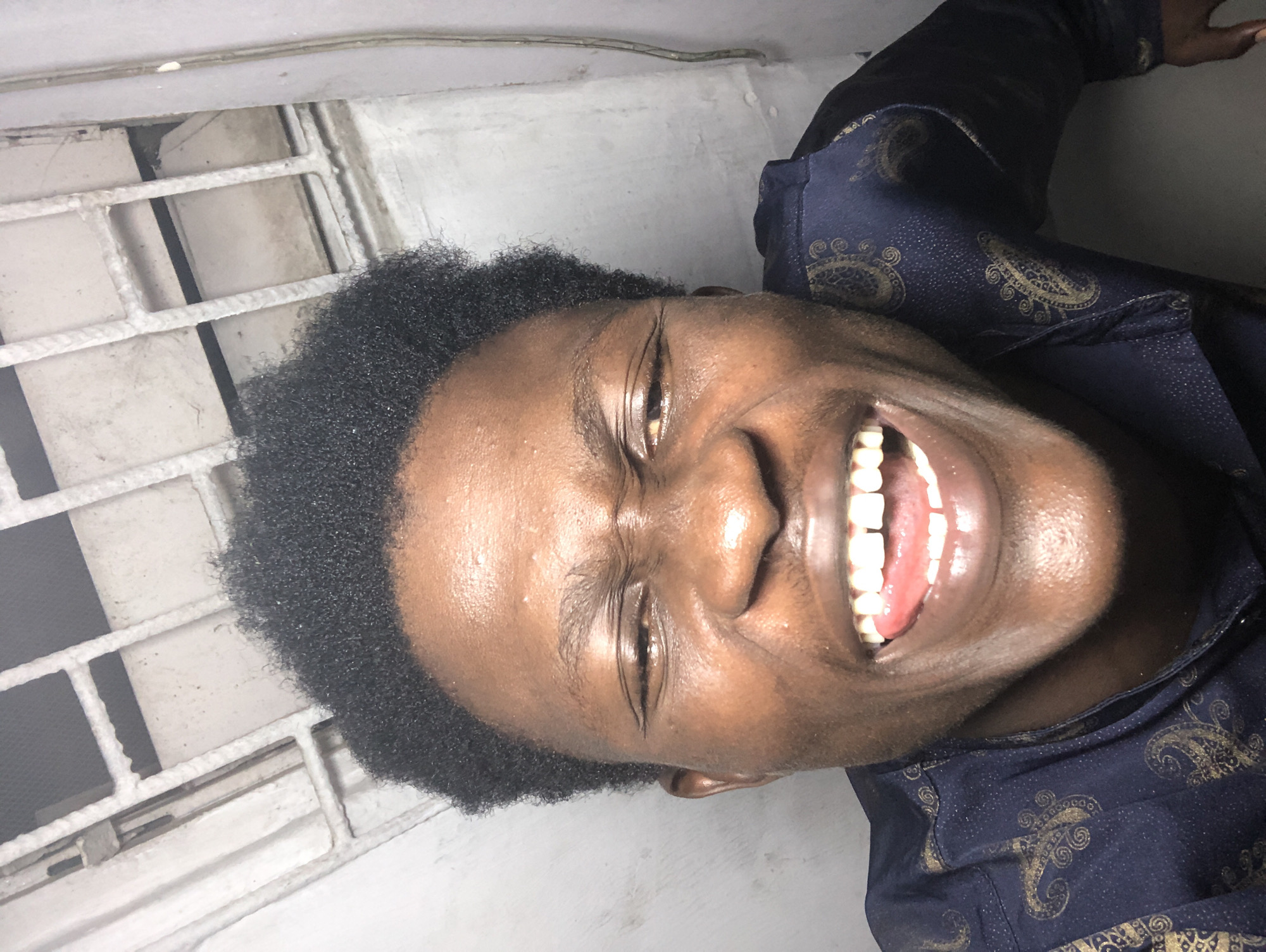 The image is a close-up, low-angle selfie of a Black man with short, dark, curly hair. He is outdoors, with a white, barred ceiling or structure visible in the upper portion of the frame. The man's face is contorted in a wide, open-mouthed laugh, with his tongue visible and his eyes squinted shut. He is wearing a dark blue shirt with a gold paisley pattern. The background walls appear to be concrete. The scene suggests an informal setting, possibly a residential area or a public space in Ado, Nigeria. The lighting is bright, creating highlights on his skin.